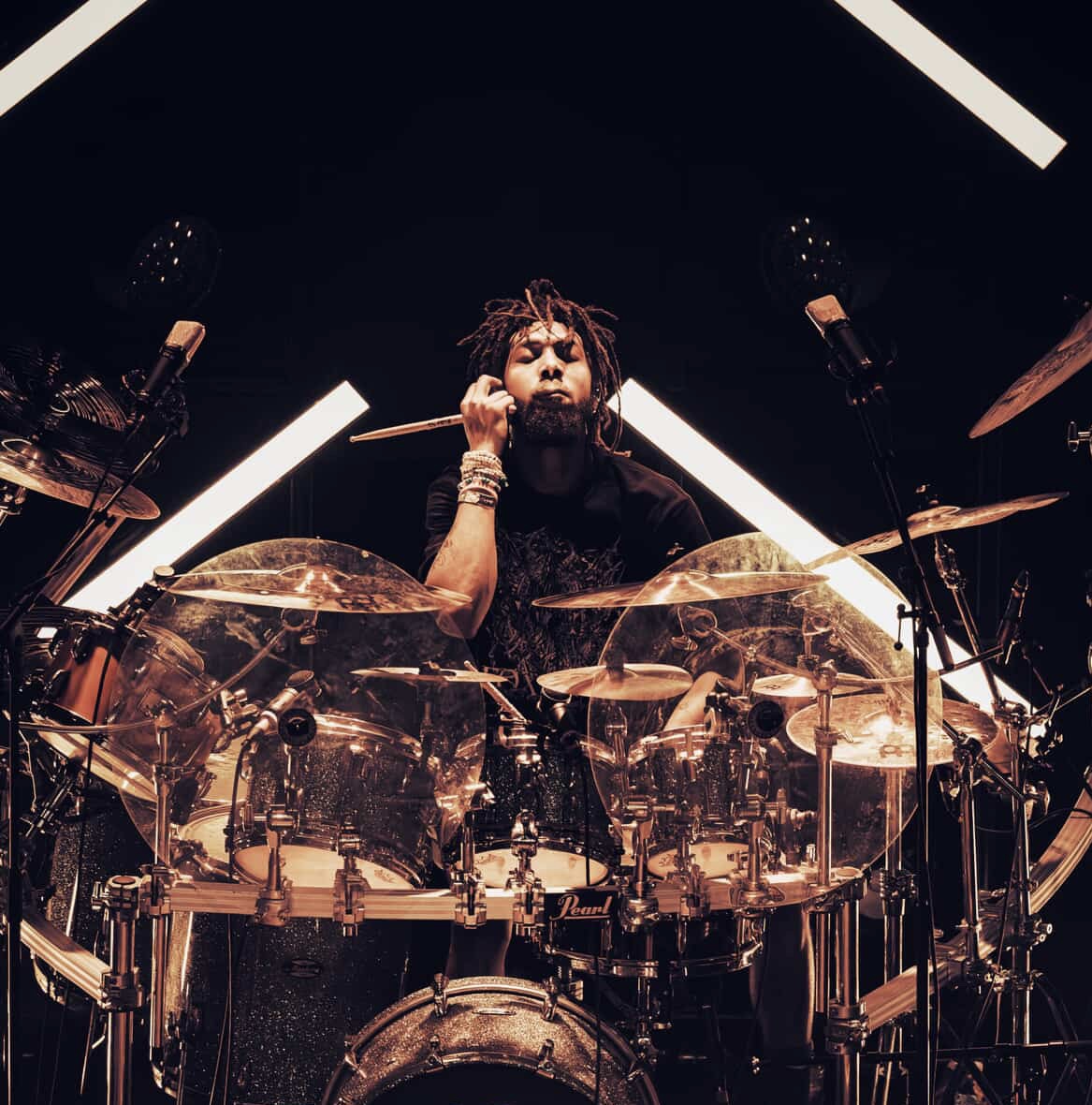 Person playing drums on stage with lighting and musical equipment, facing forward, with dark background.