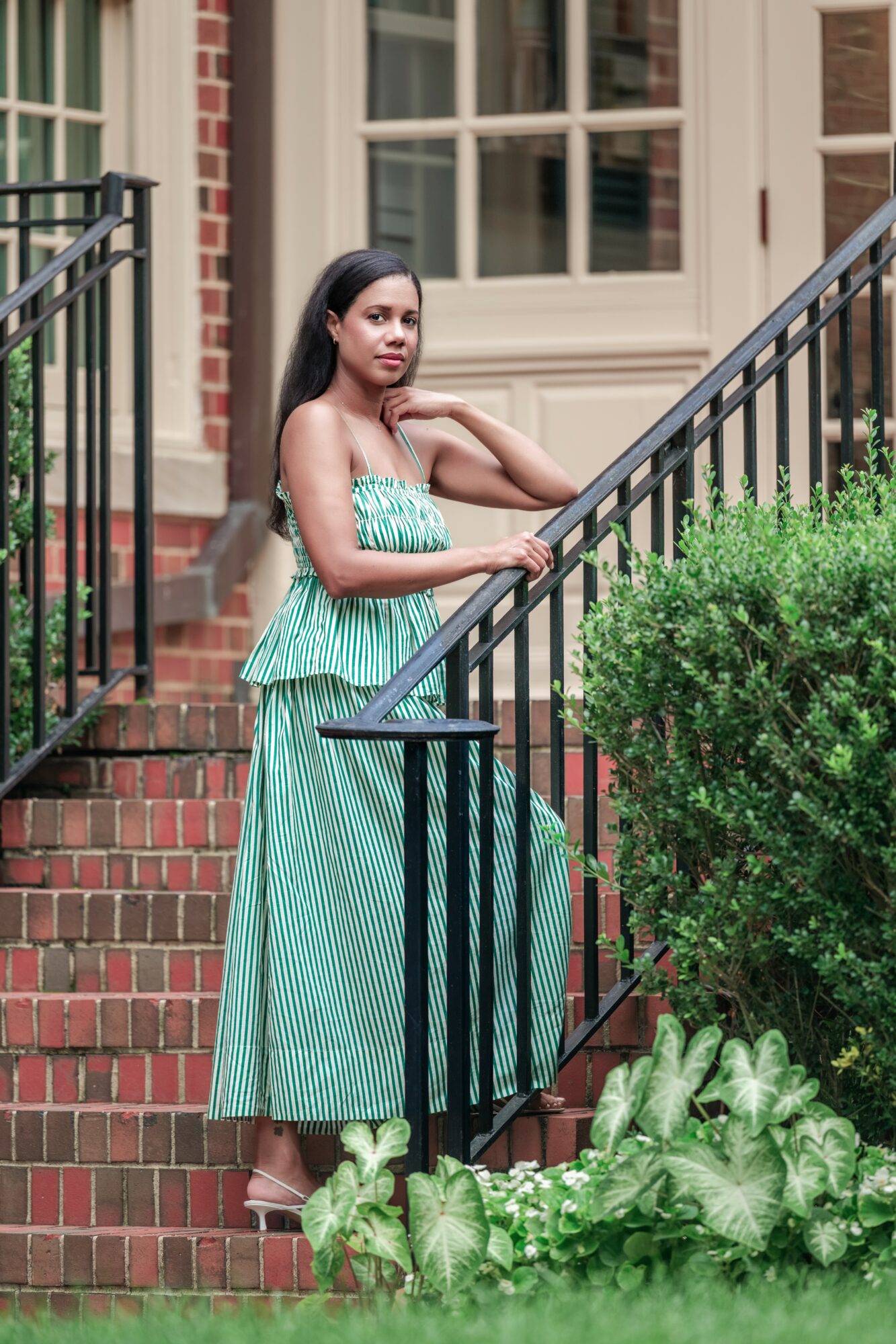 Young woman in a green strapless dress standing on brick stairs outside a building, with black railing and greenery nearby.
