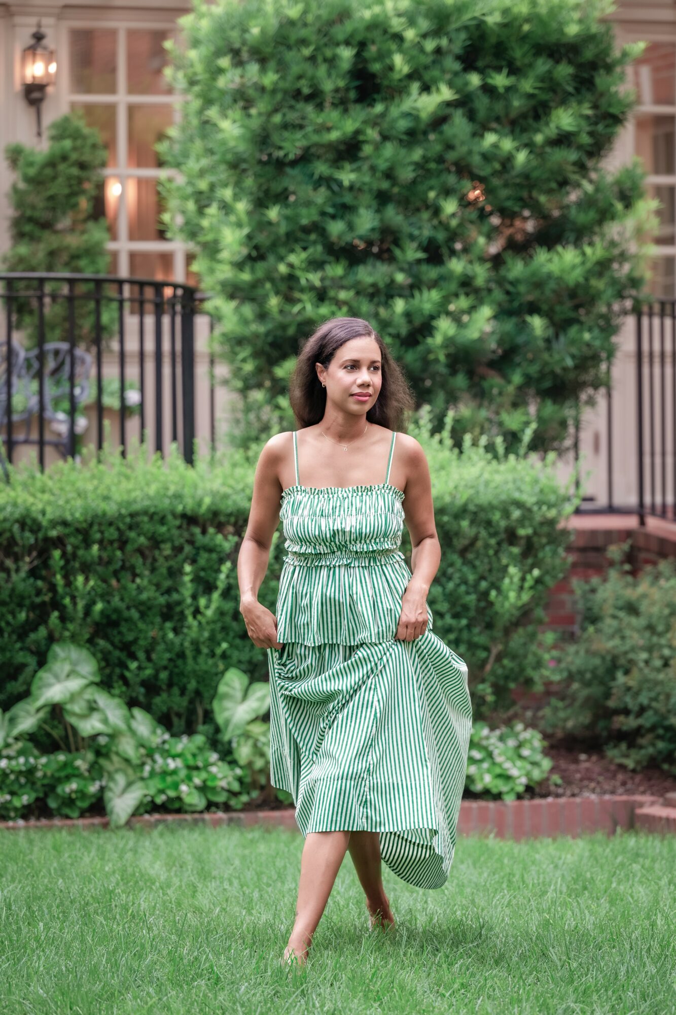 Young woman in a green and white striped dress walking on grass with trees and a fence in background.