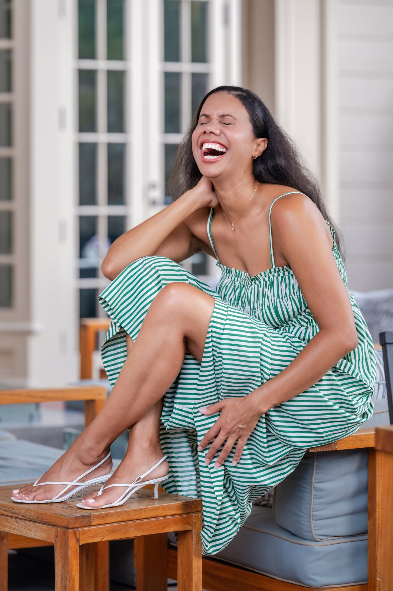 Woman laughing outdoors, sitting on a wooden bench, wearing a striped dress and clear heels, with a house and windows in background.