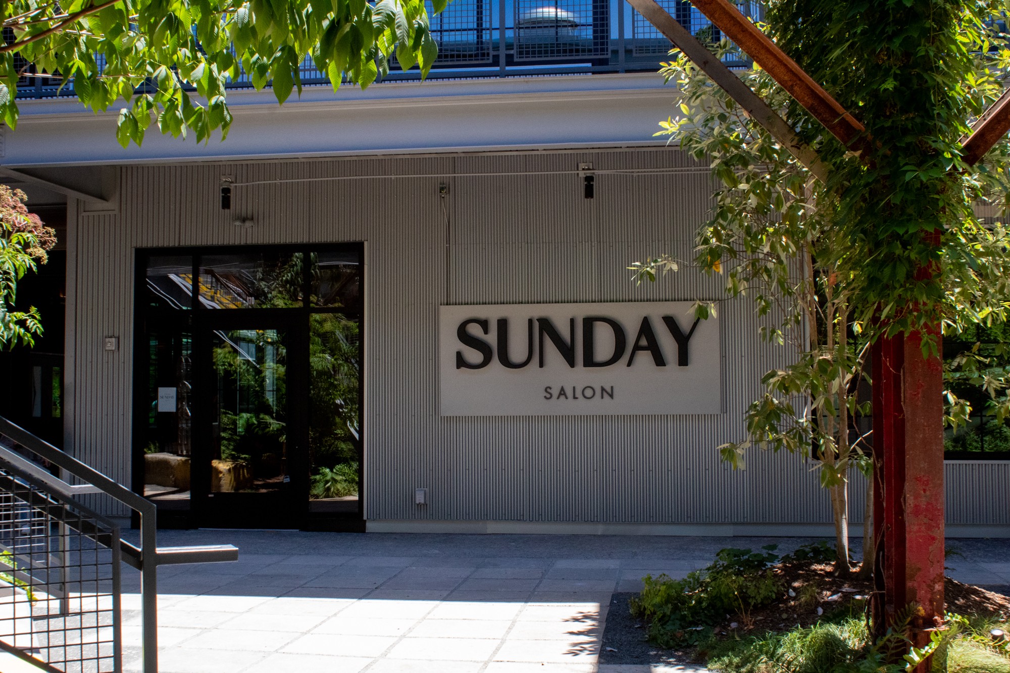 Facade of a salon with a sign reading 'SUNDAY SALON' and glass doors, surrounded by trees and greenery.
