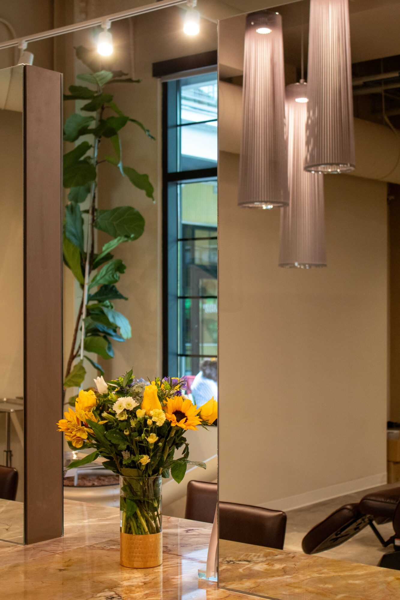 Interior space with hanging pendant lights, a tall window, a large plant, and a vase of yellow flowers on a table.