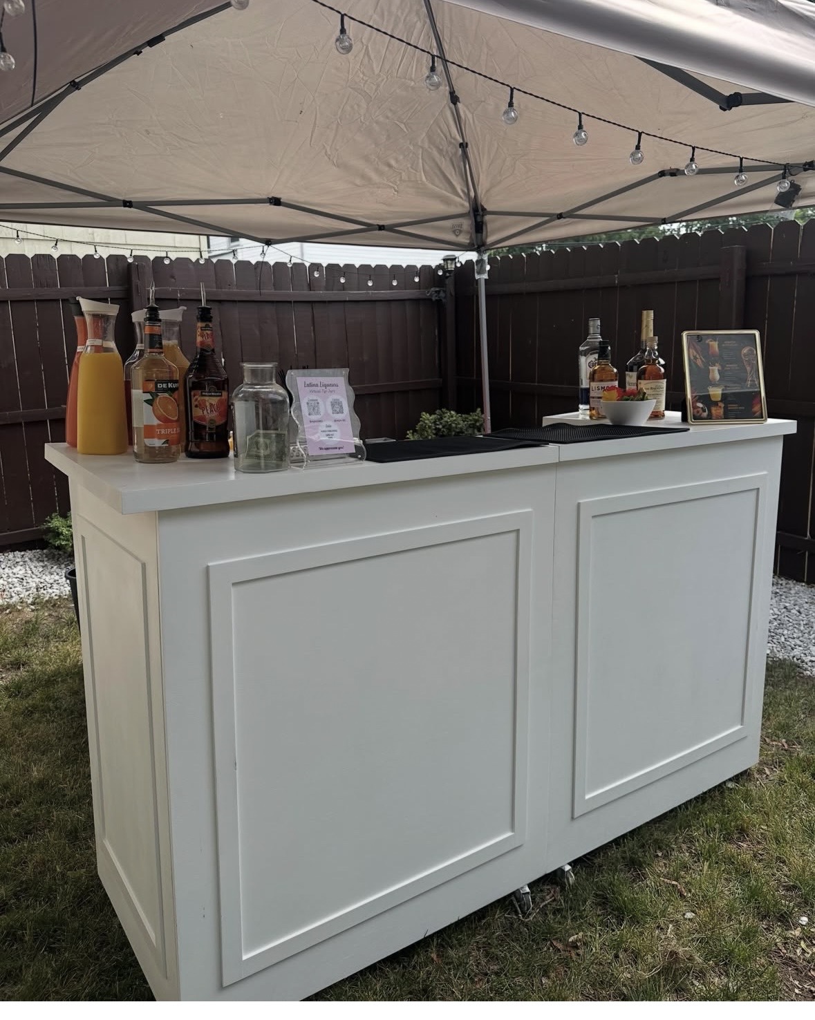 White outdoor bar with bottles and glasses under a canopy, fenced yard background.