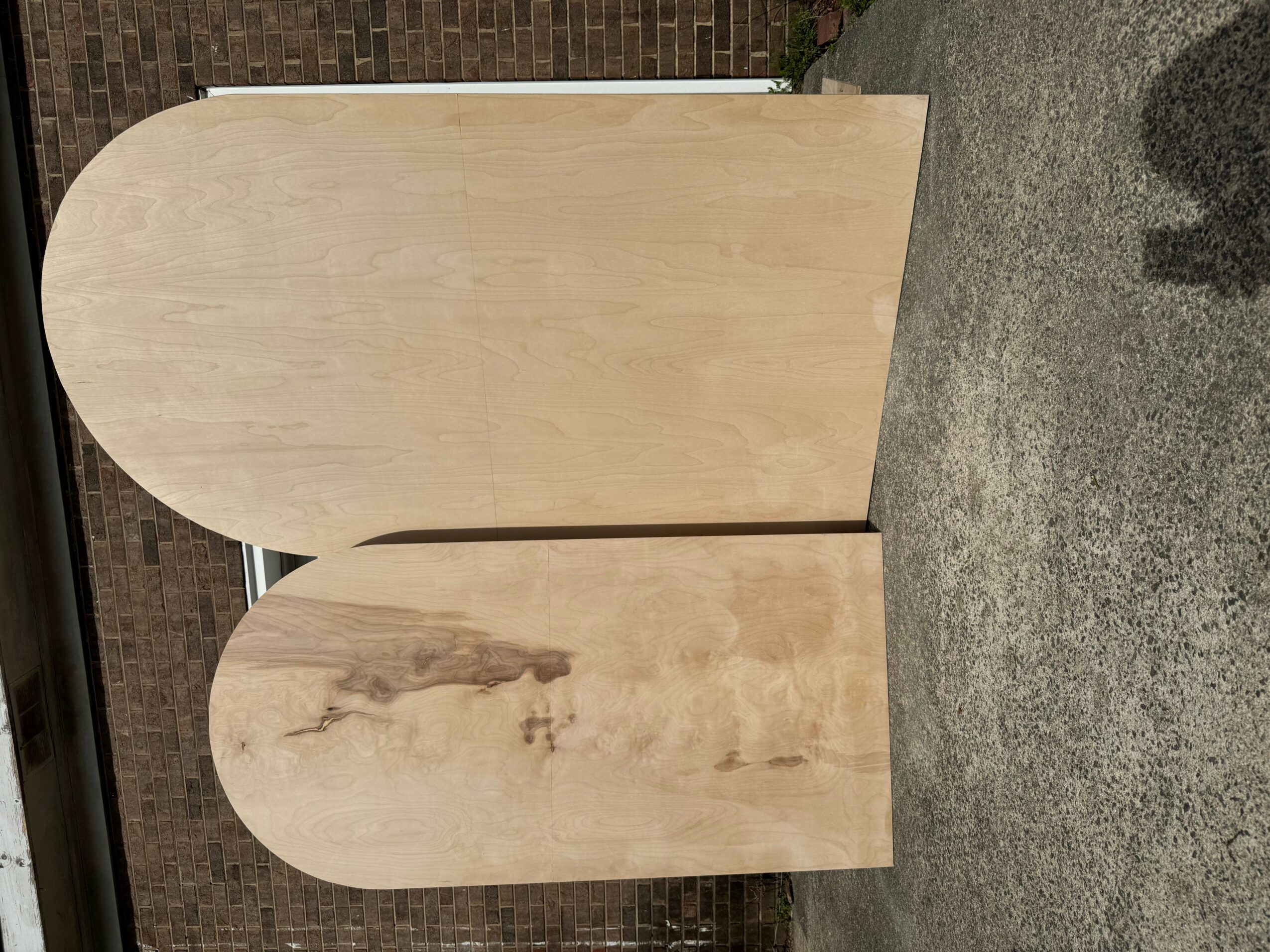 Two wooden panels with rounded tops leaning against a brick wall, on a concrete floor.