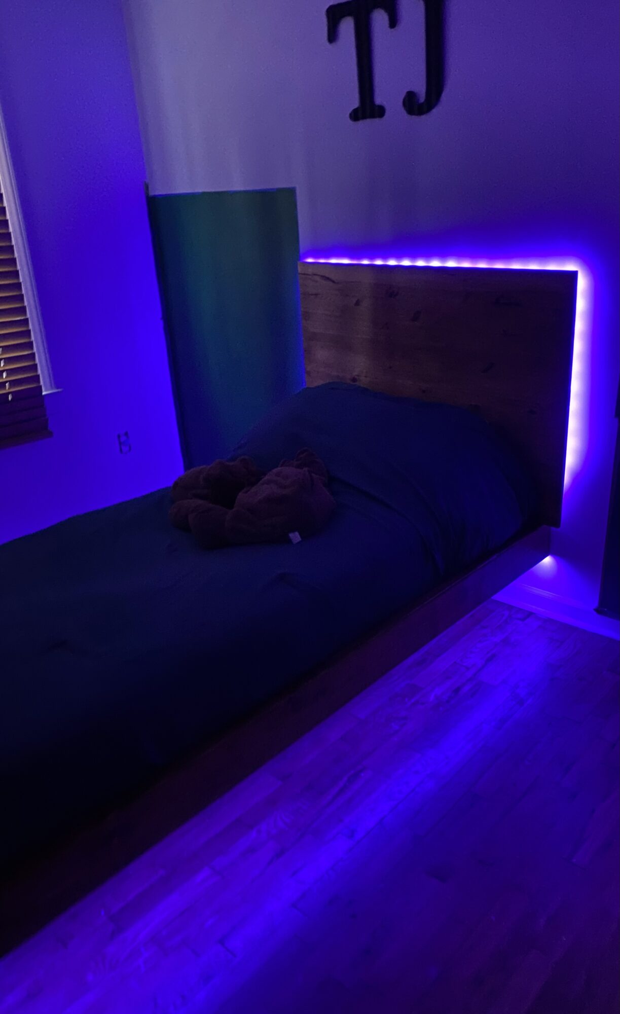 Bed with dark bedding and a stuffed animal, illuminated by purple LED lights, in a dimly lit room.