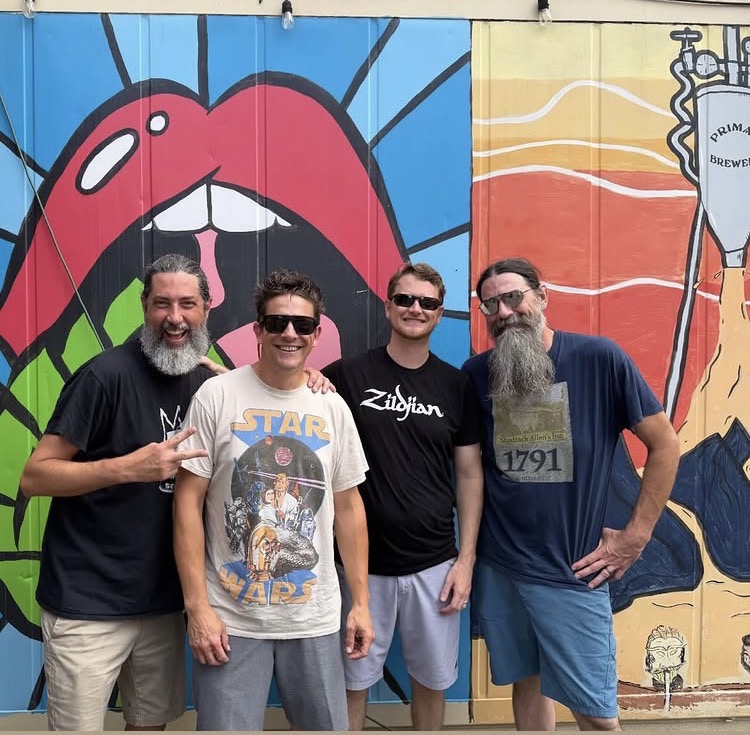 Four people stand in front of colorful mural with lips, mountains, and a beer glass, smiling at the camera.