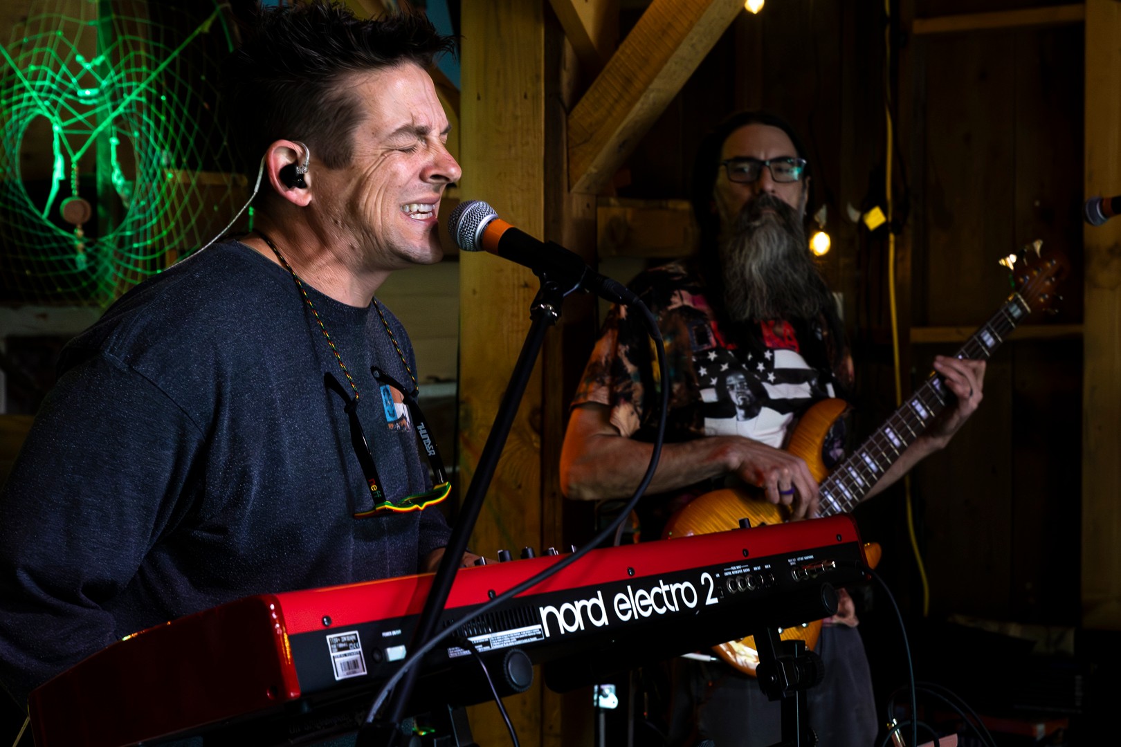 Two musicians perform on stage, one singing into a microphone and playing a keyboard, the other playing guitar with a beard and glasses.