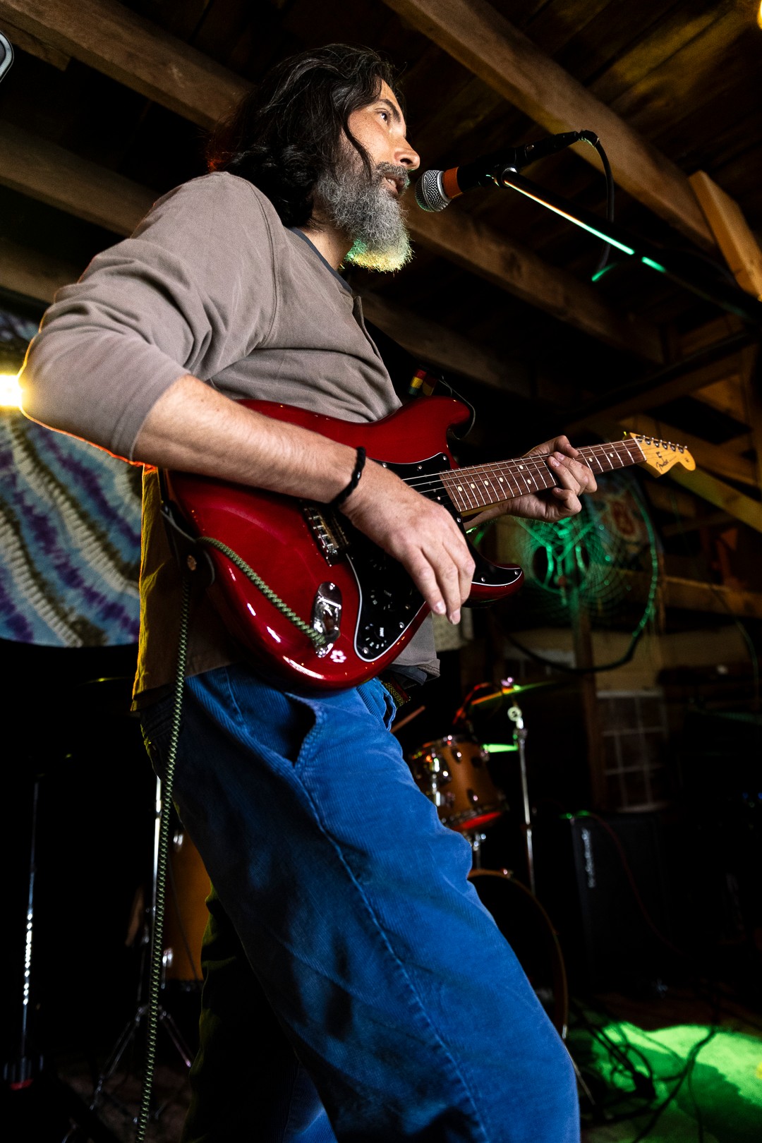 Person playing red electric guitar on stage with microphone, wooden ceiling, and musical equipment in background.