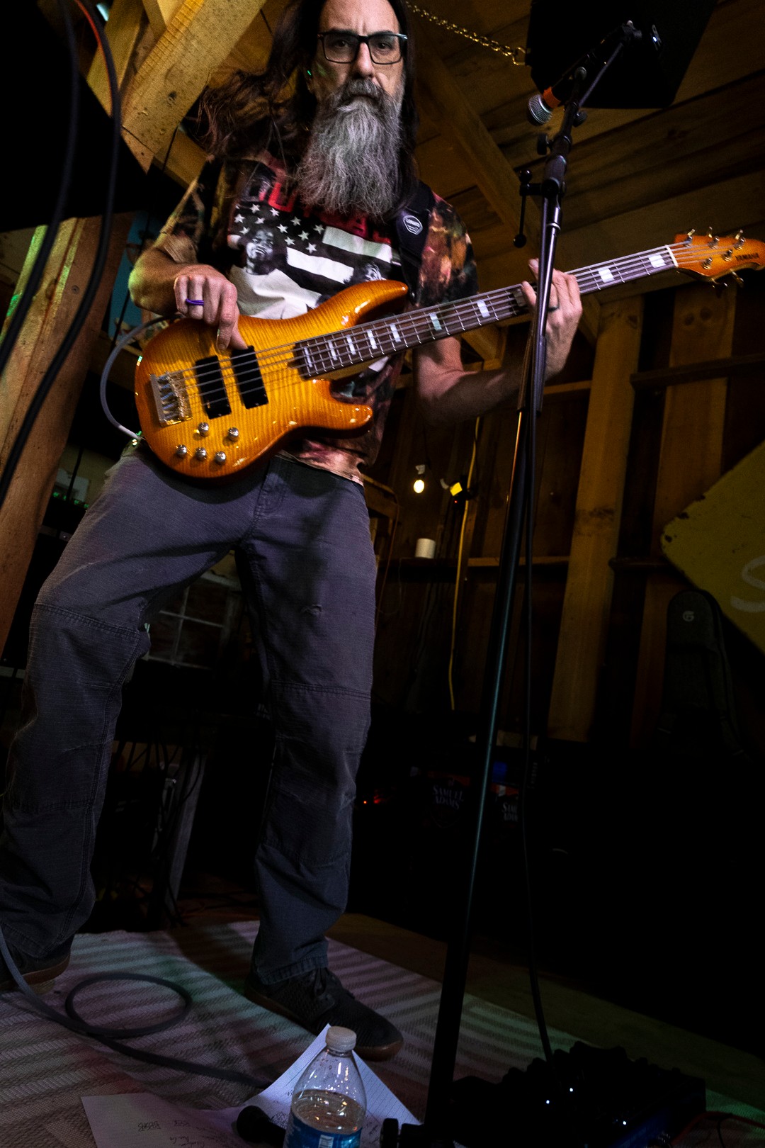 Man with long hair and beard playing an orange electric bass guitar on stage, with wooden background and microphone stand.
