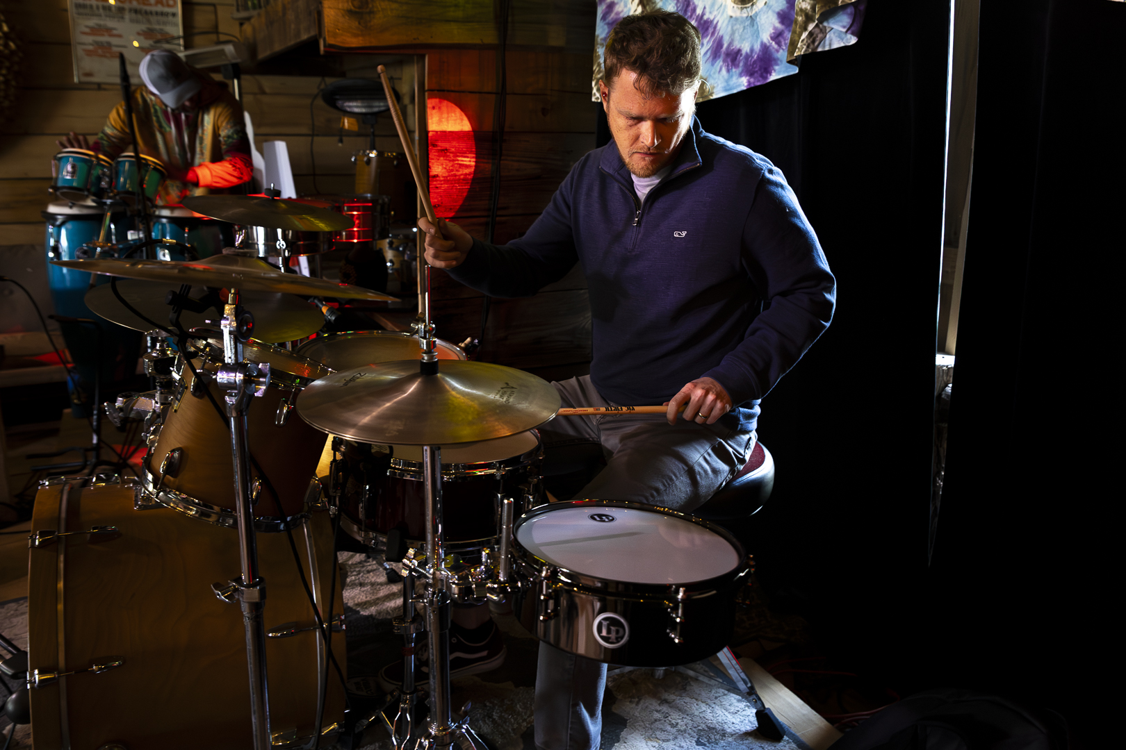 Man playing drums in a dimly lit room with colorful background lights, focused on his instrument.