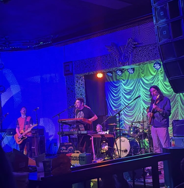Band performing on stage with colorful lighting and draped backdrop, three musicians playing instruments, one singing into microphone.