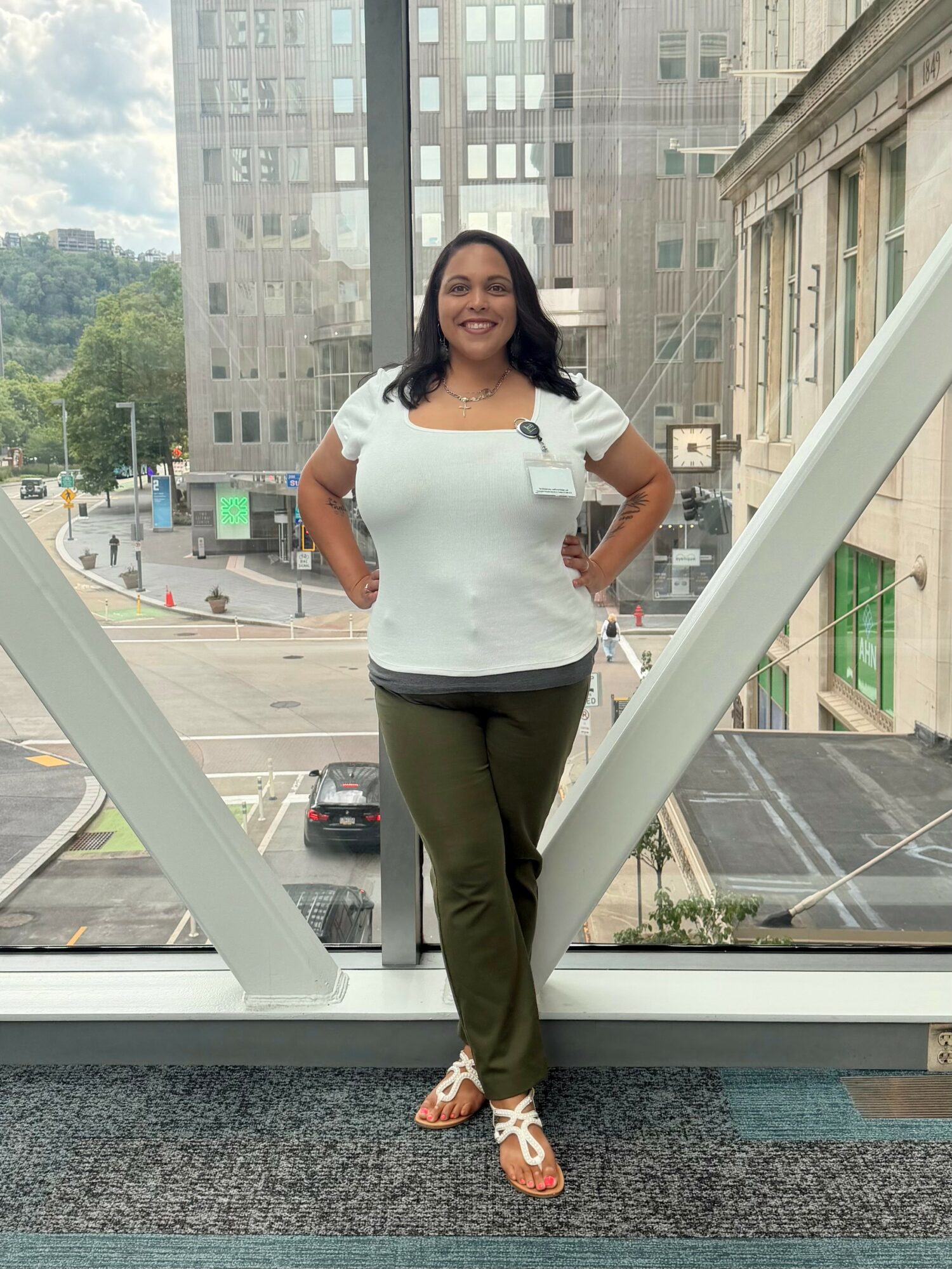 Woman standing in front of large window with cityscape view, smiling, hands on hips, wearing white top and green pants.