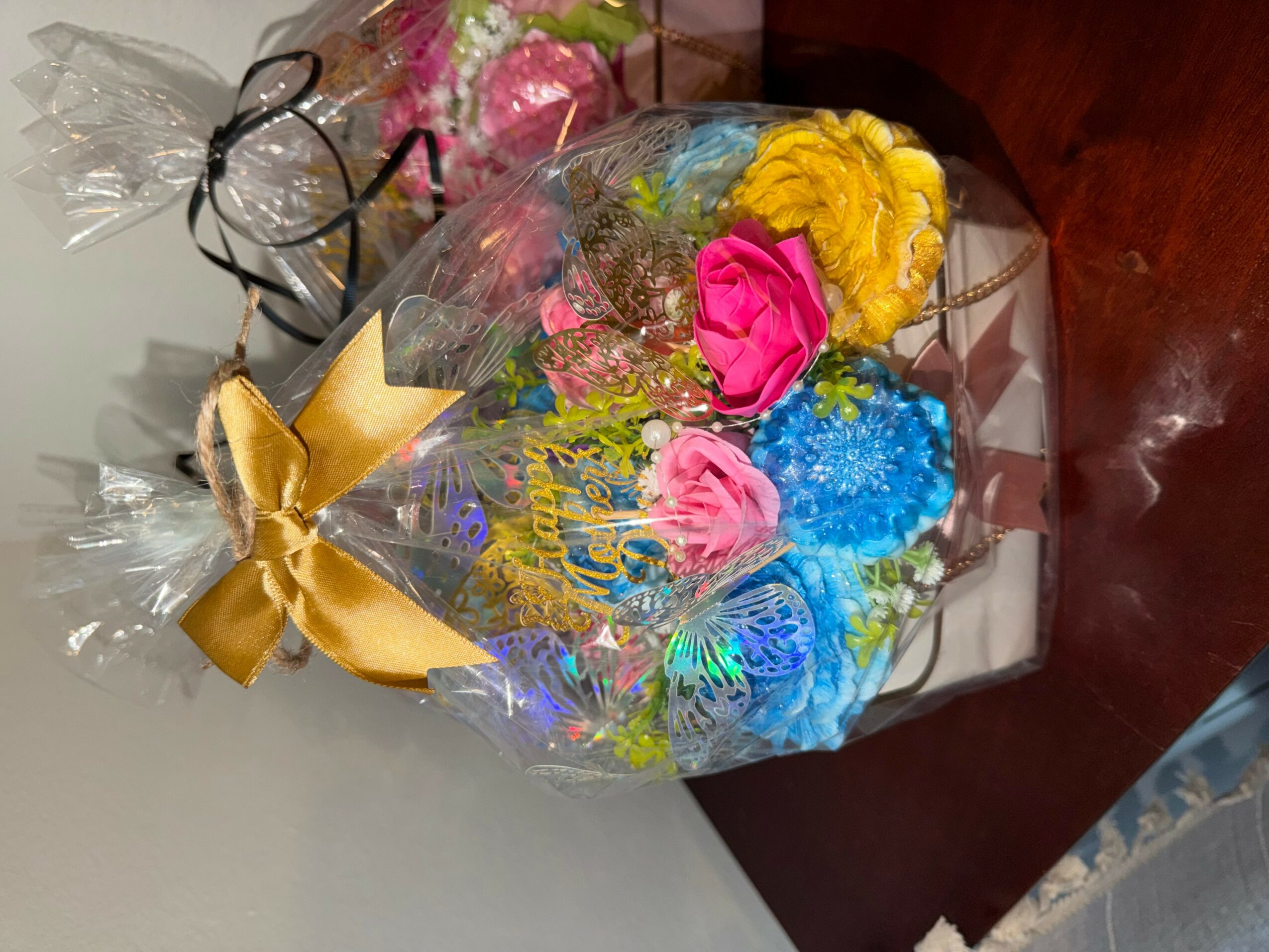 Colorful bouquet of artificial flowers wrapped in clear plastic with a gold ribbon, placed on a wooden surface.