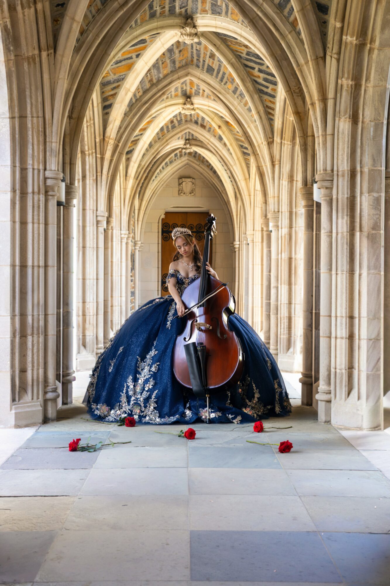 Woman in a blue gown playing a cello in a Gothic-style arched corridor with scattered roses on the floor.