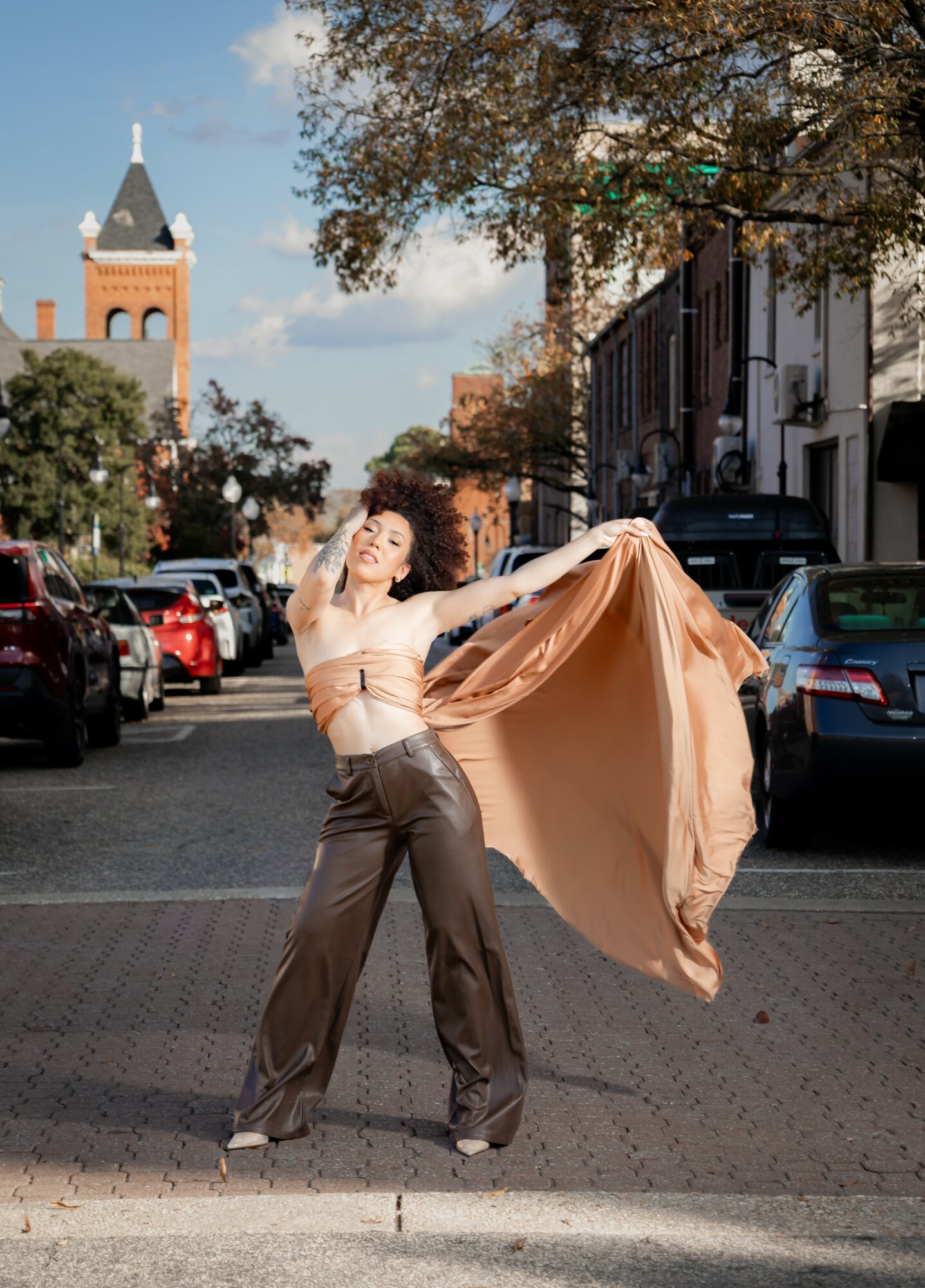 Woman dancing outdoors on a city street, holding a flowing fabric, with trees and buildings in the background.