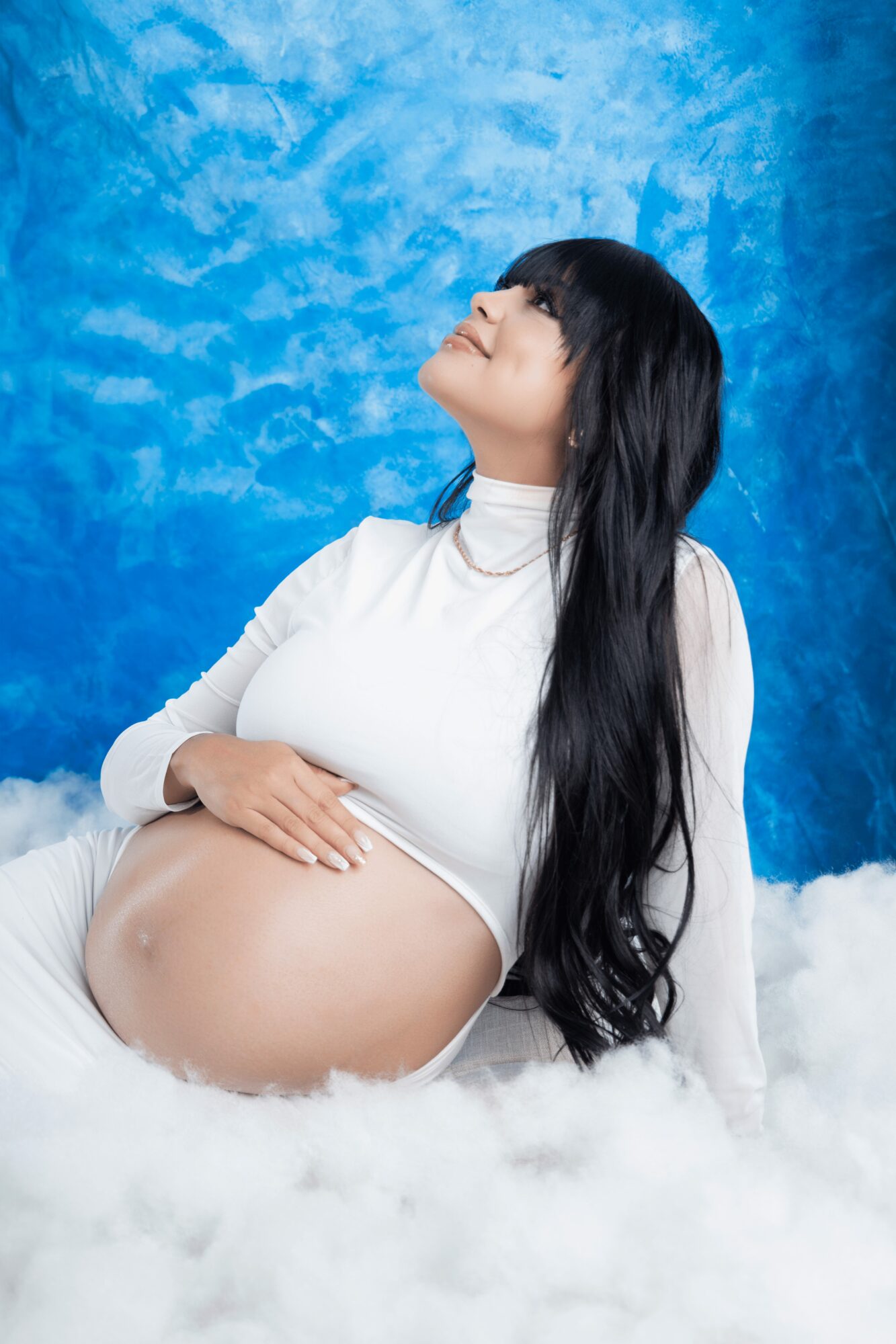 Pregnant woman with long black hair, wearing a white top, smiling with eyes closed, against a blue background, sitting on white fluffy material.