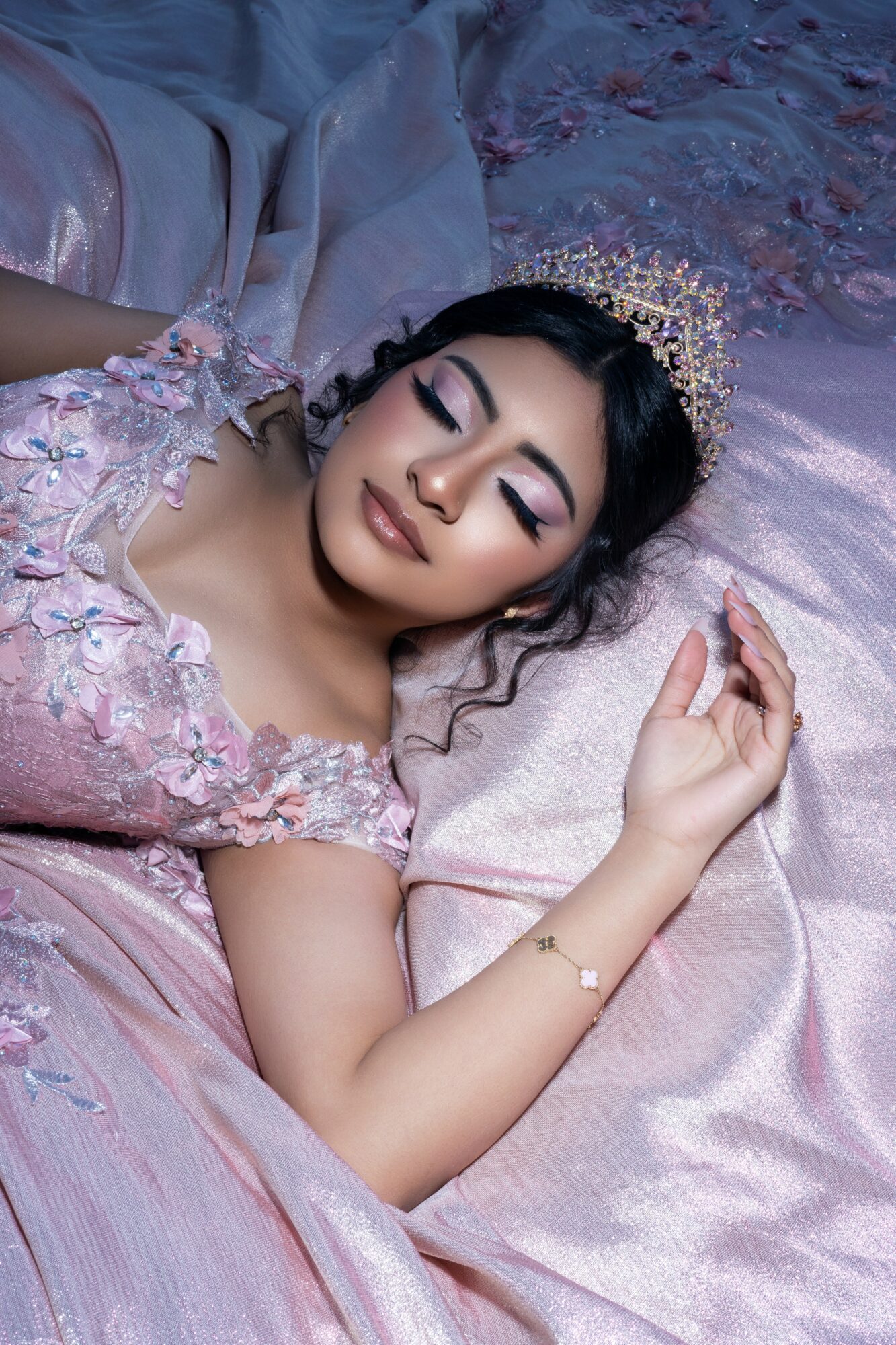 Woman with makeup and a crown lying on a bed with pink and purple bedding, eyes closed, relaxed pose.
