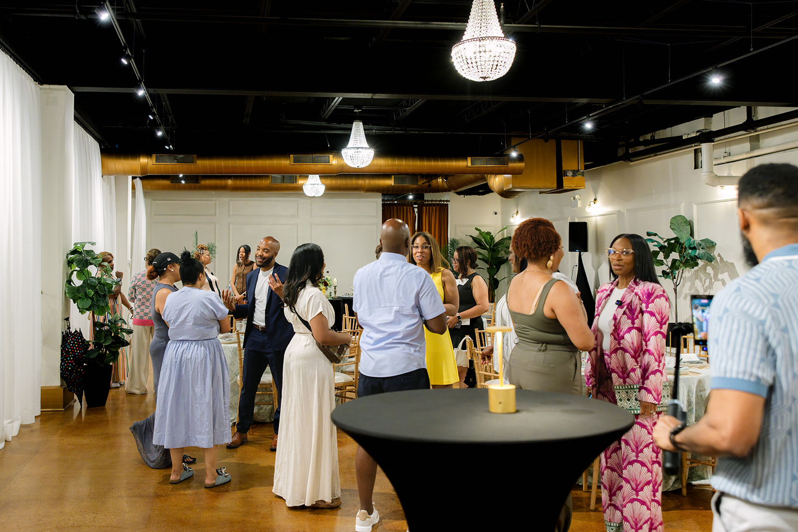 People gathered in a decorated indoor space with plants, tables, and hanging lights, engaging in conversation and socializing.