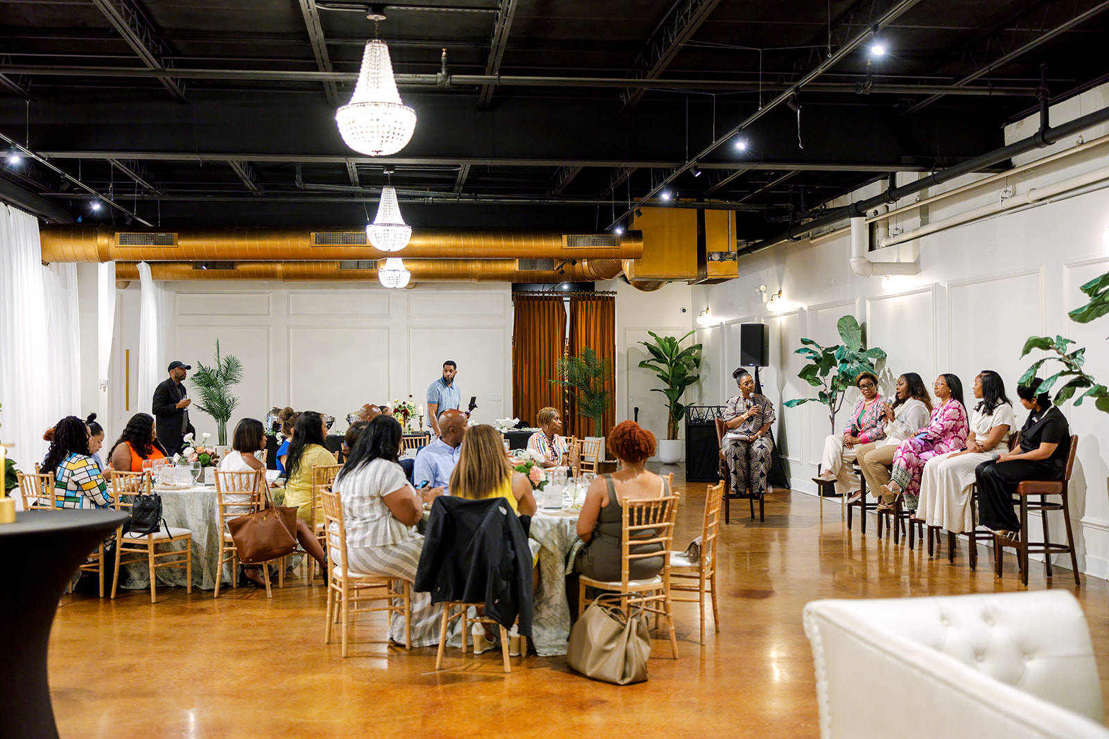 People seated at tables in a spacious room with plants, chandeliers, and a speaker at the front.