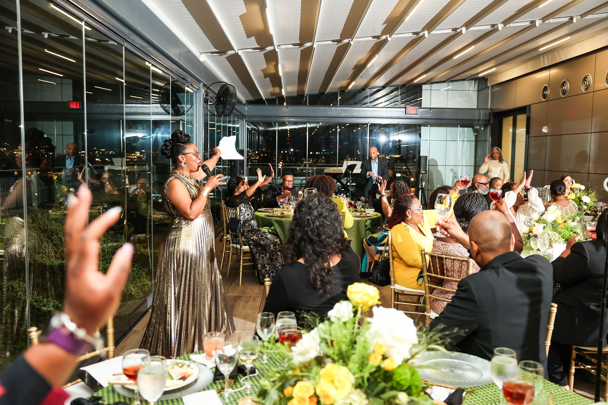 Woman in a metallic dress speaking to seated guests at a formal event in a modern venue with large windows and floral arrangements.