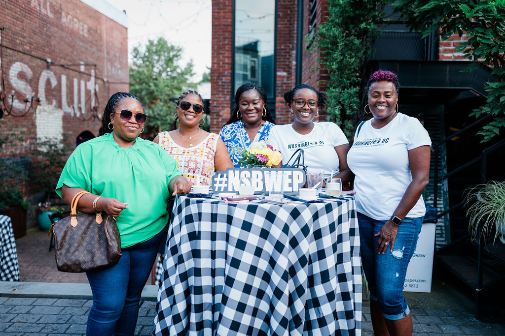 Five women standing outdoors behind a table with a checkered tablecloth, smiling, with a cake and a sign reading '#NSBWEP'.