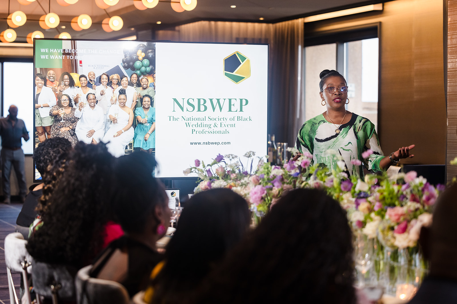 Woman speaking at a conference with a large screen displaying NSBWEP logo and group photo behind her, audience seated in front.