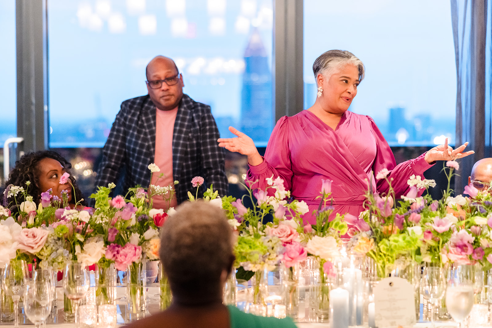 Older woman in pink dress speaking at a table with flowers, two men and a girl listening, large window in background