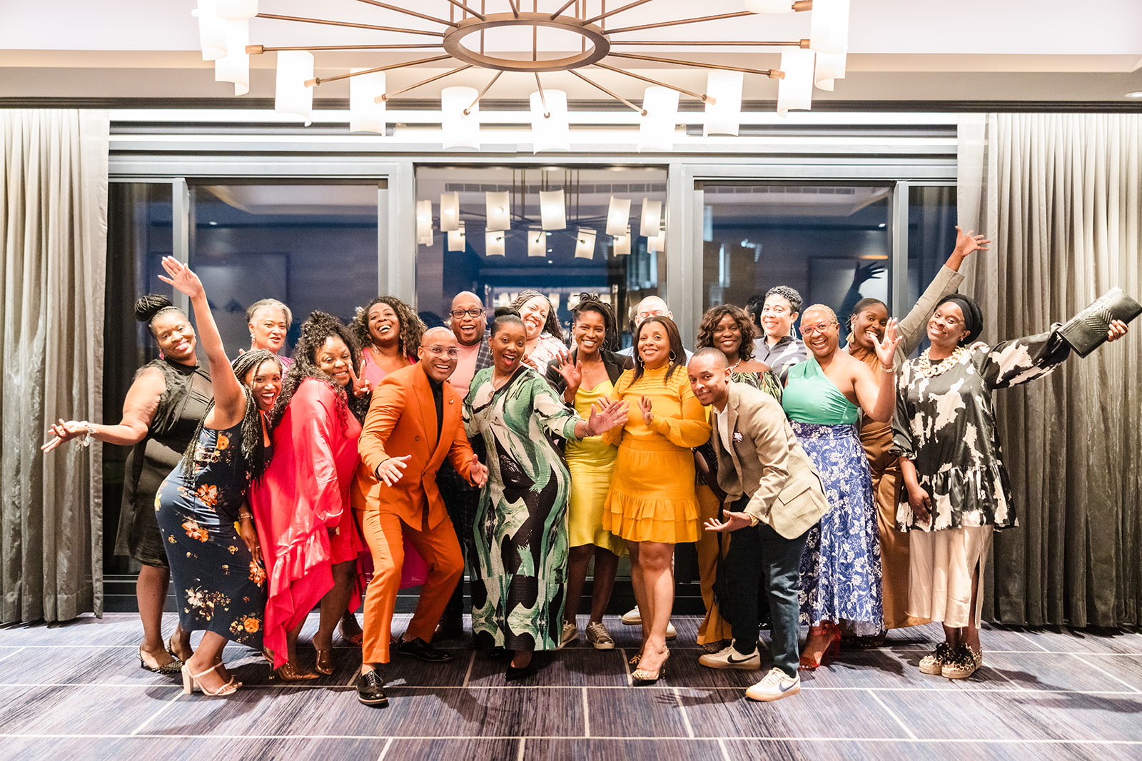 Group of diverse women posing together indoors, smiling and celebrating, with some raising arms and others making gestures.