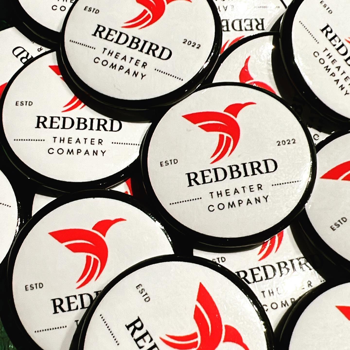 Multiple round tokens with Redbird Theater Company logo, featuring a red bird and text, scattered on a surface.