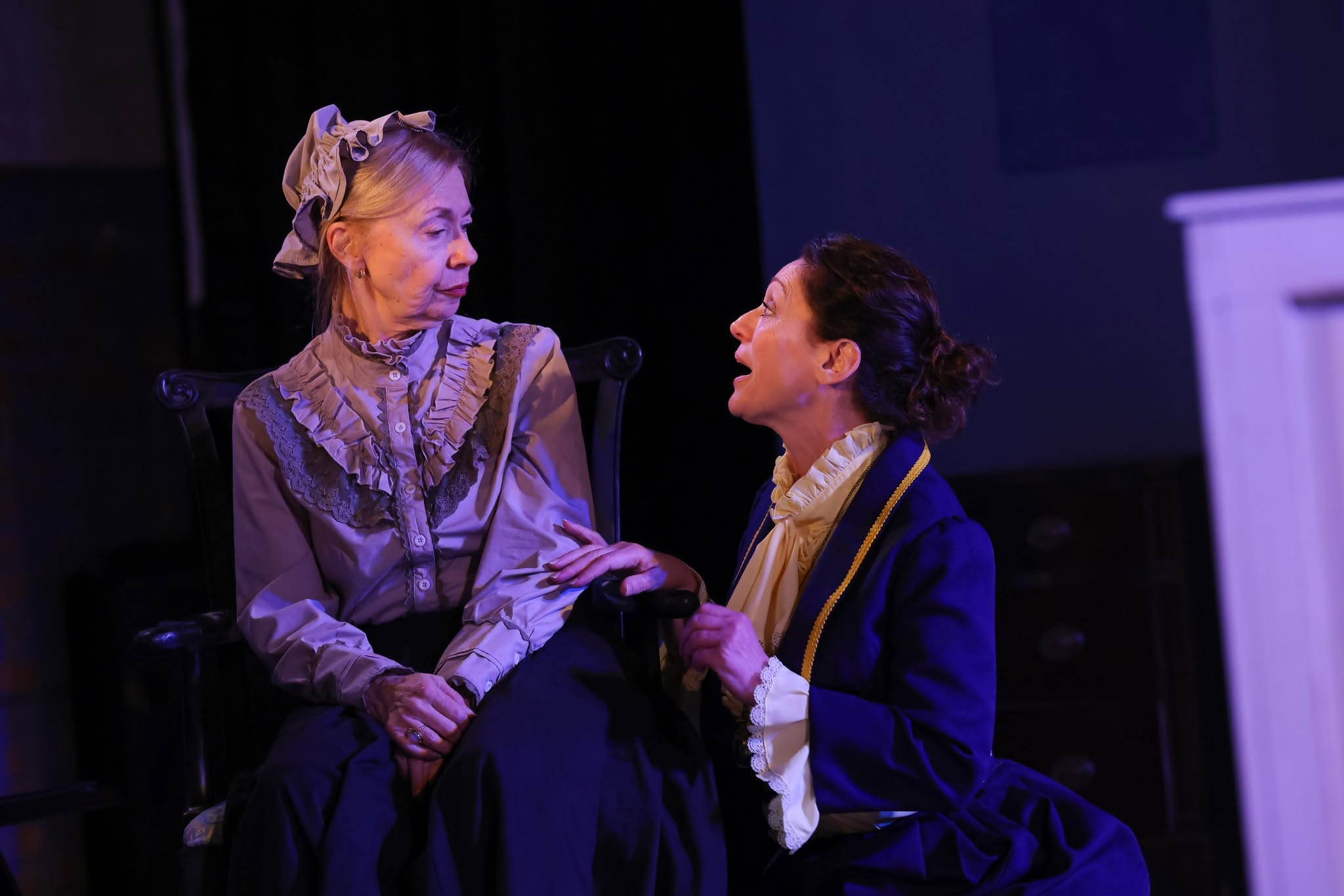 Two women in period costumes sit and converse on stage, one older and one younger, with dark background and stage lighting.