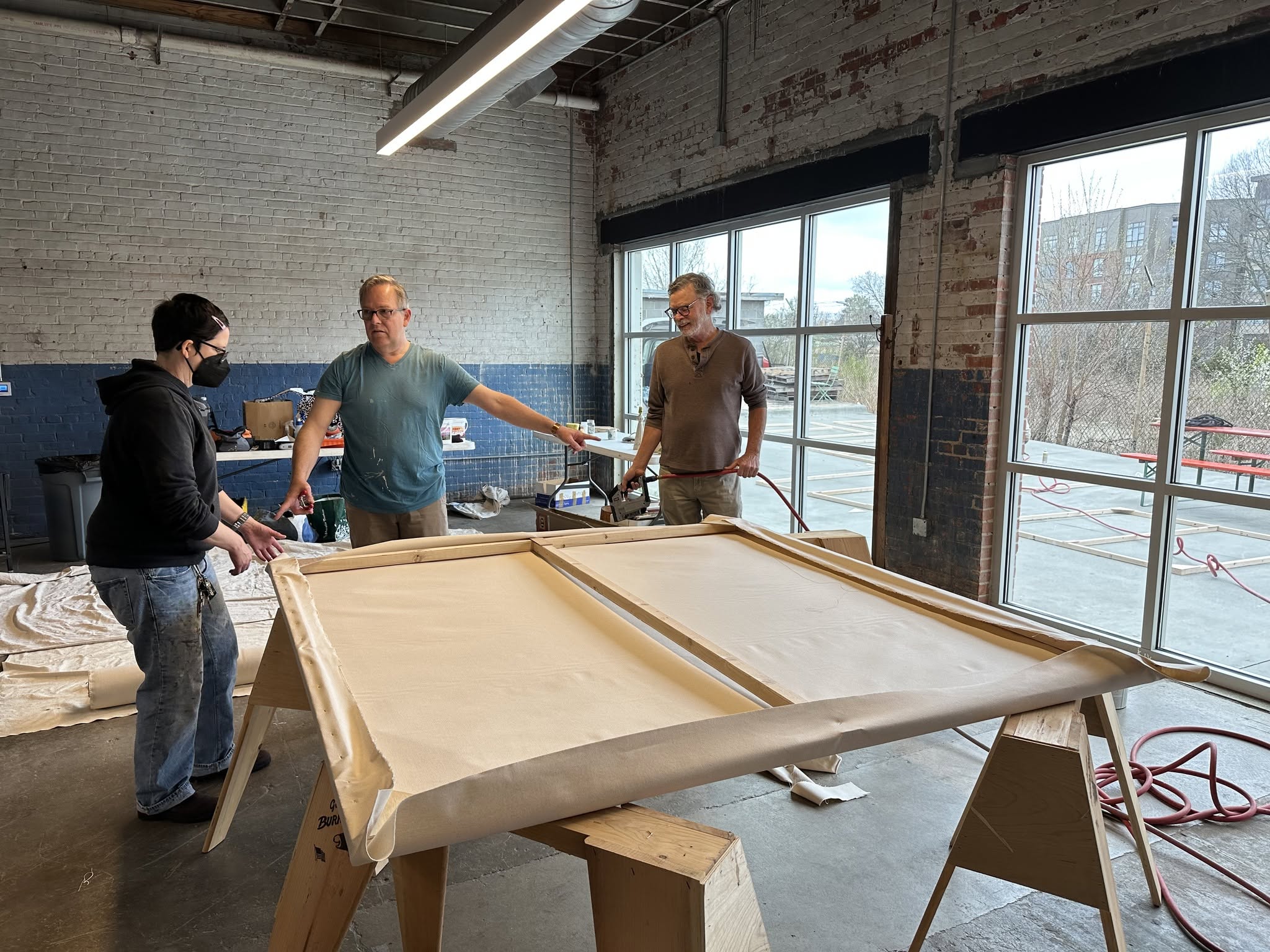 Three men in a workshop around a large wooden frame on a table, with large windows and brick walls in the background.