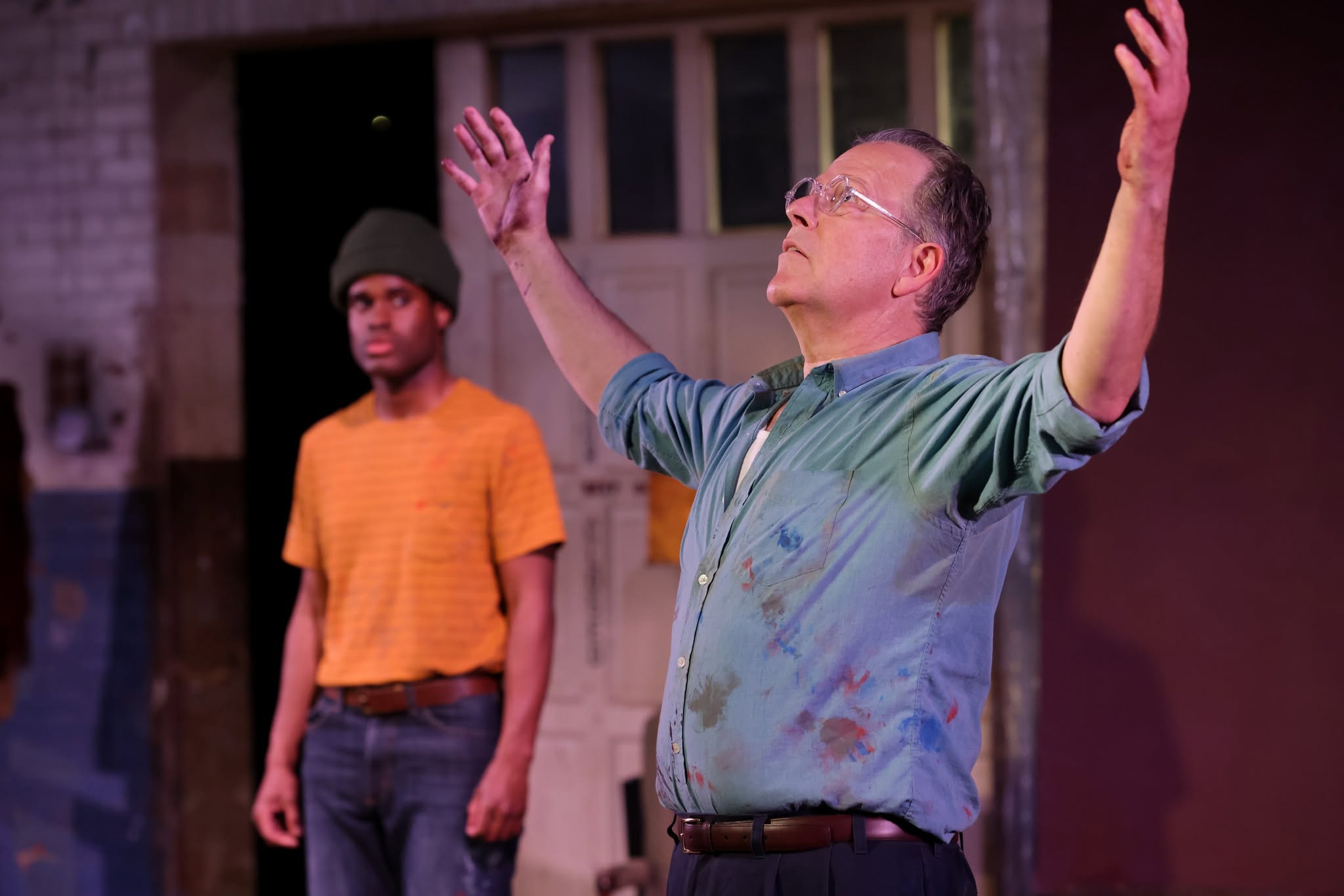 An older man with glasses and a colorful shirt raises his arms outdoors, with a young person in an orange shirt and beanie in the background.