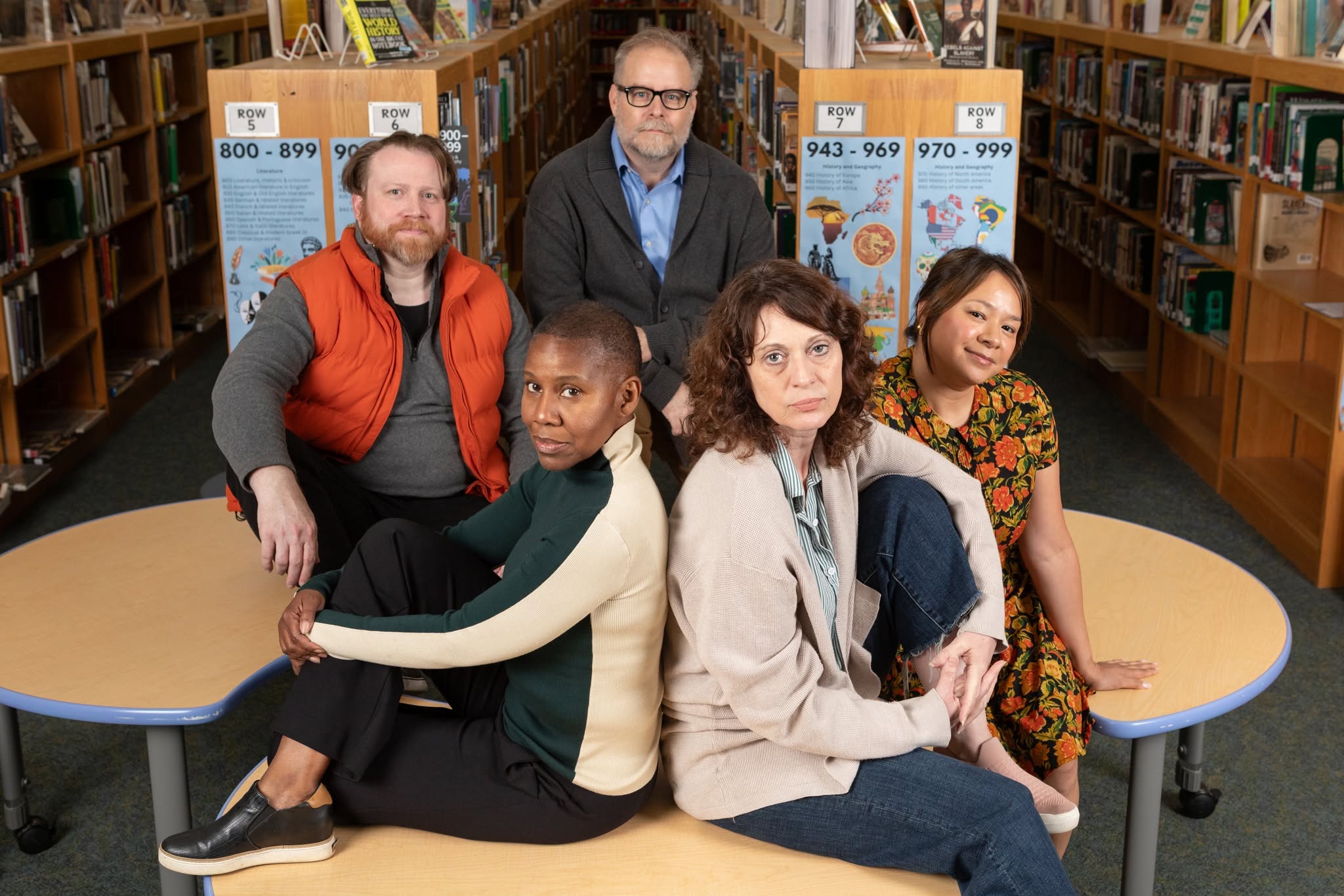 Six people sitting and standing in a library, with bookshelves and posters in the background, smiling at the camera.