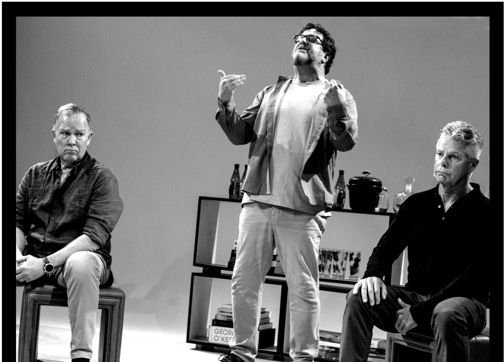 Three men on stage, one standing and speaking, two seated on either side, with a plain background and furniture.