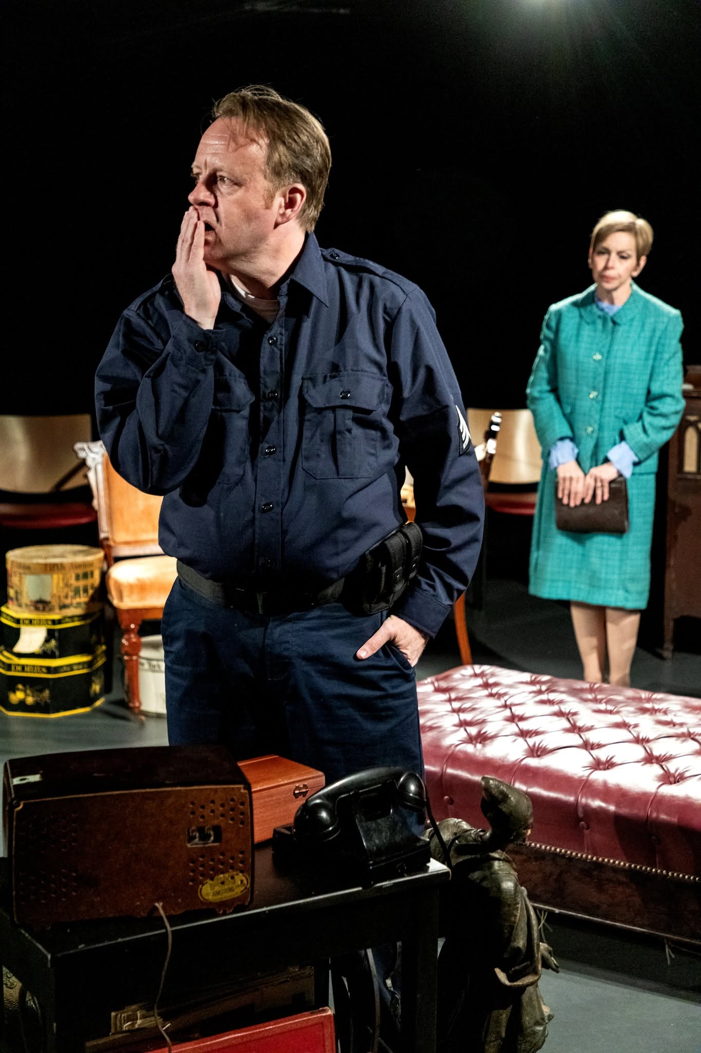Man in dark jacket with hand on face, woman in teal coat in background, stage setting with furniture and props