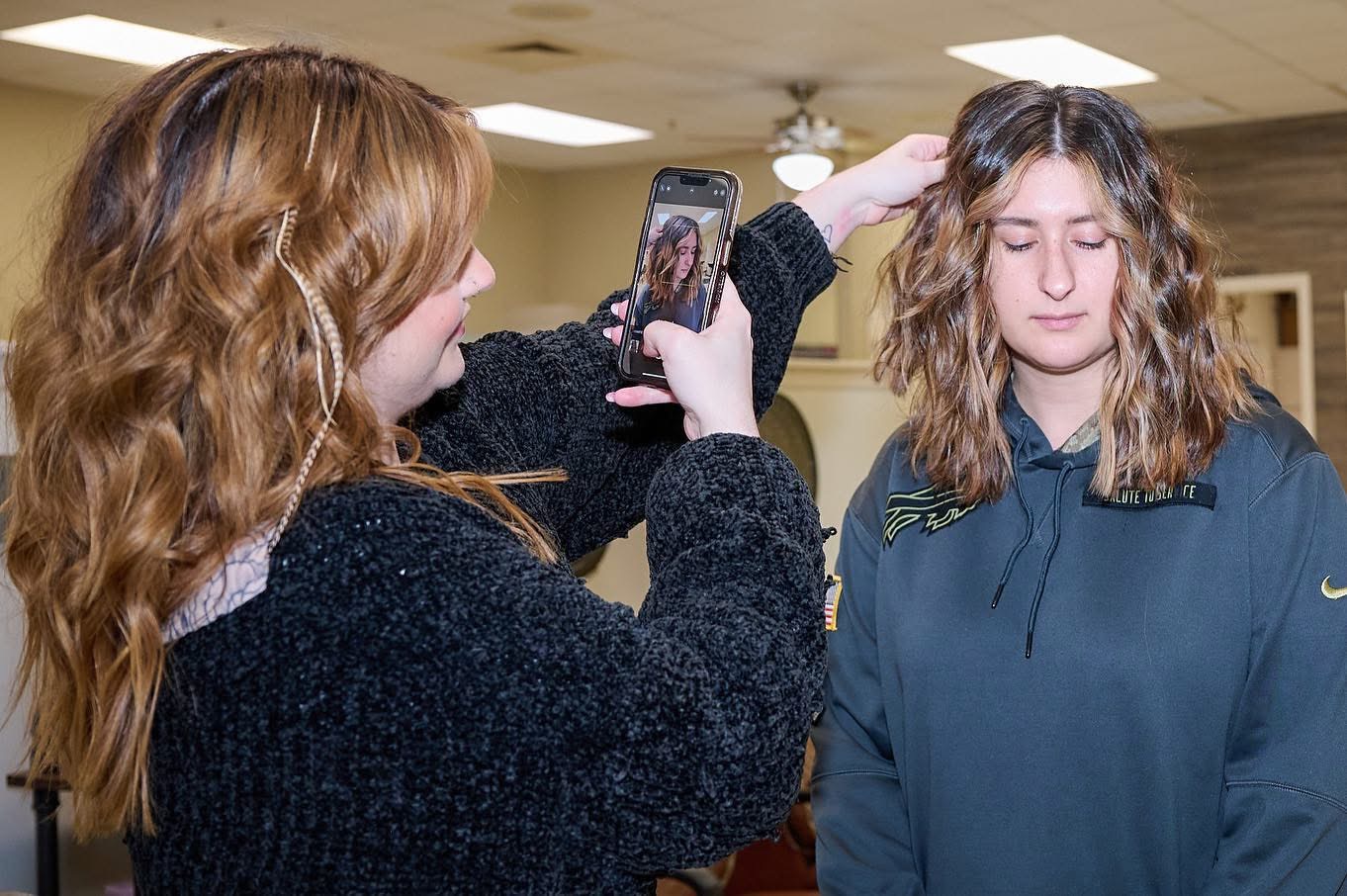 Person taking a photo of another person with a smartphone indoors, both with wavy hair, one with eyes closed.