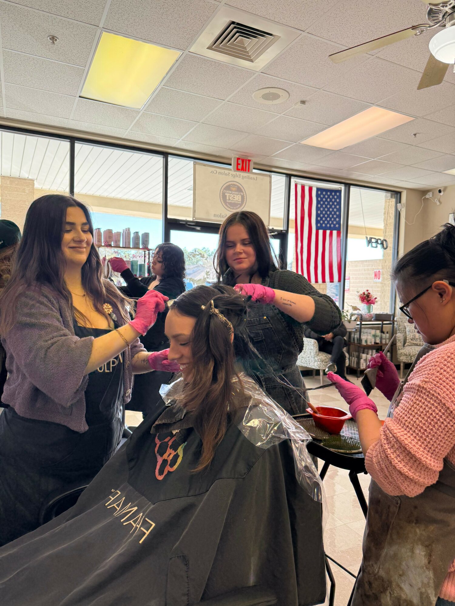 People in a hair salon getting hair treatments, with an American flag and cityscape visible through the window.