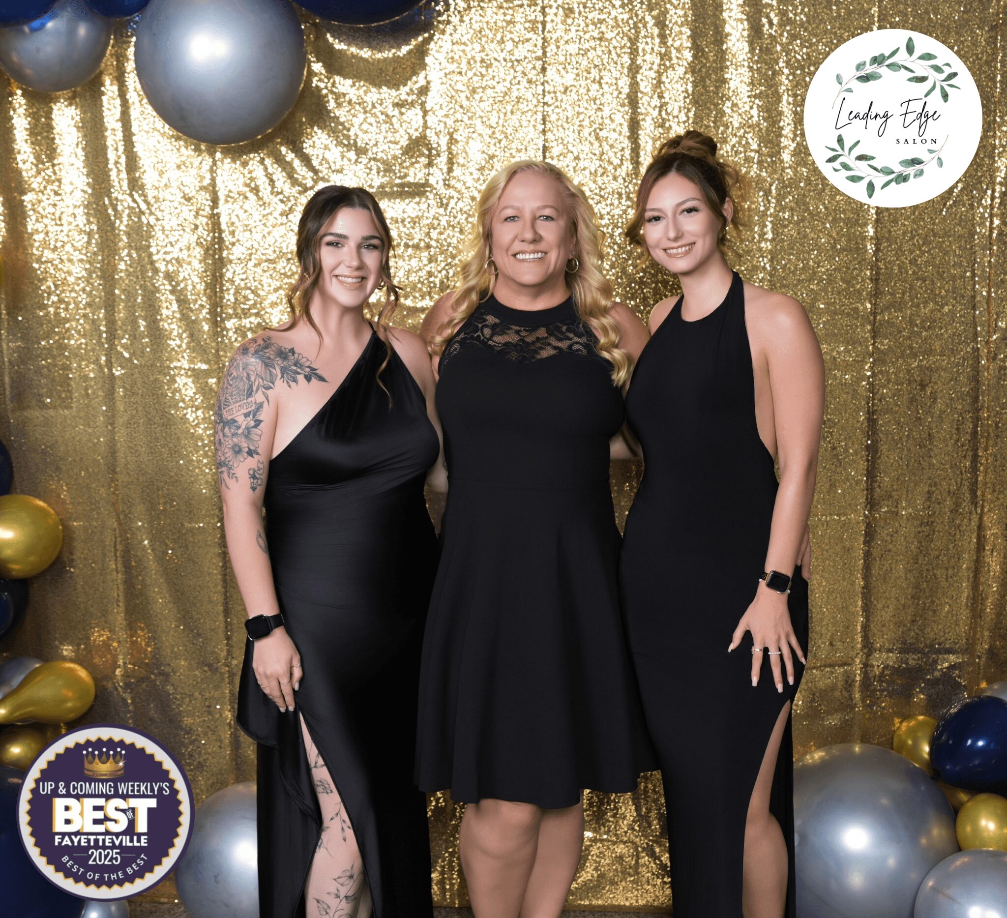 Three women in black dresses standing in front of a gold backdrop with balloons and a sign, smiling.