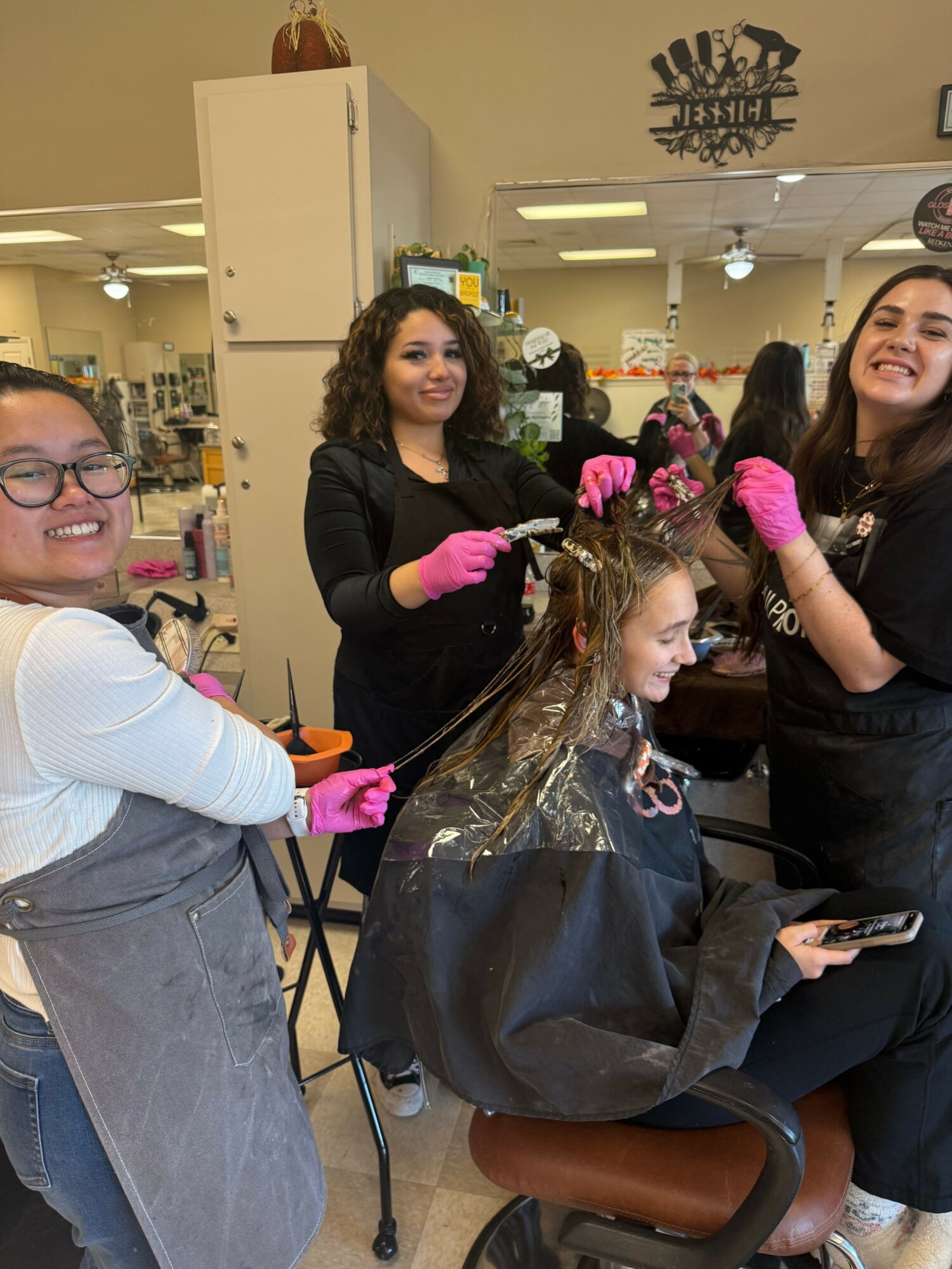 A woman with glasses and a gray vest is getting her hair styled by a hairstylist in a salon. The stylist is wearing pink gloves.
