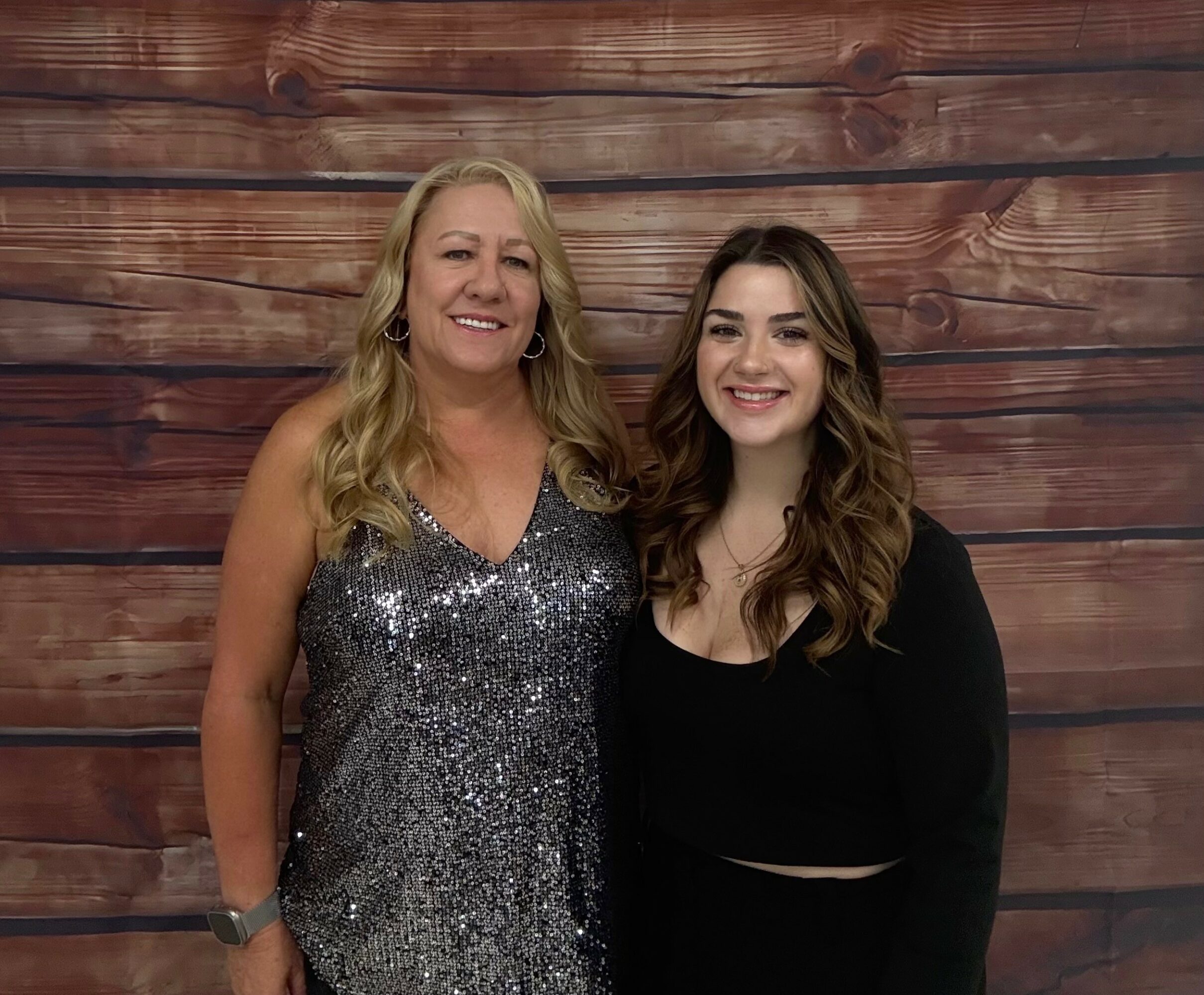 Two women standing side by side smiling, one wearing a sparkly dress and the other in a black top, against a wooden background.