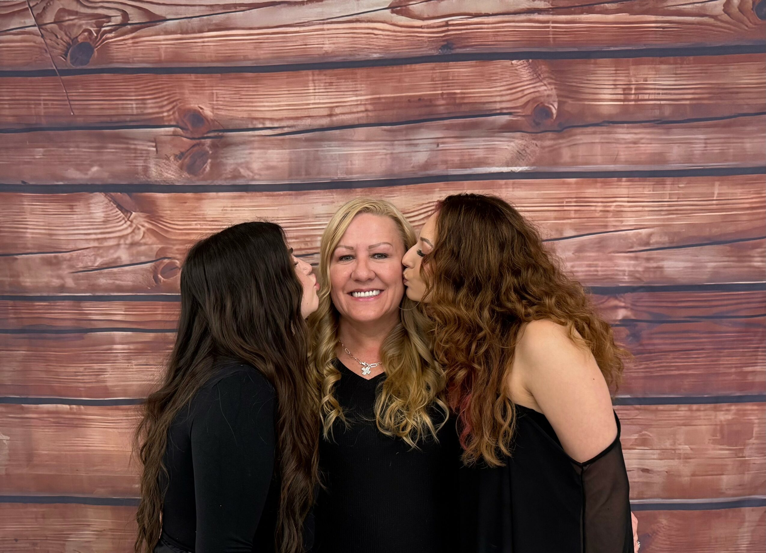 Two women kiss a smiling woman in the middle against a wooden background.