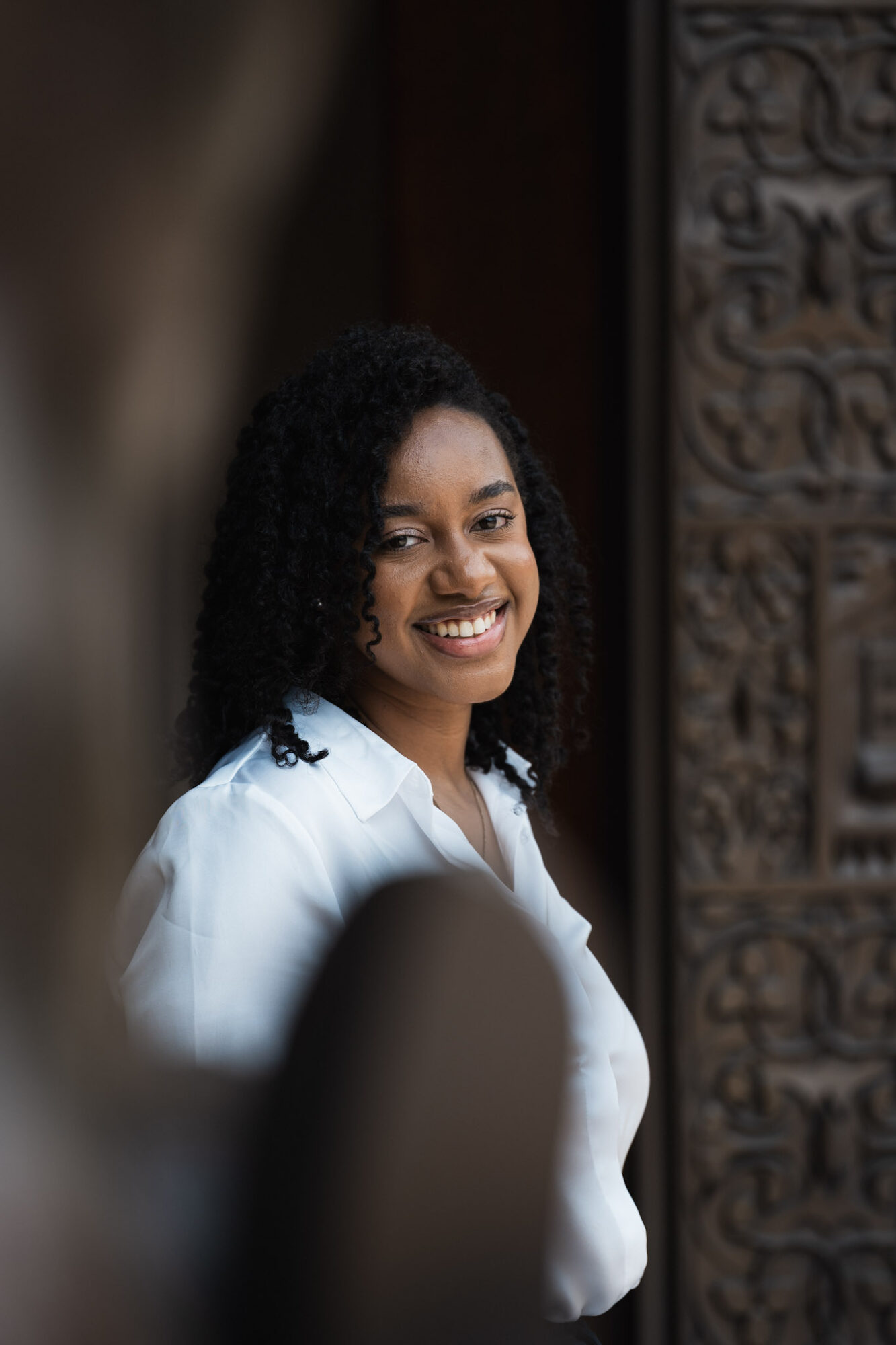 Smiling woman with curly hair wearing a white shirt, standing near a carved wooden panel.