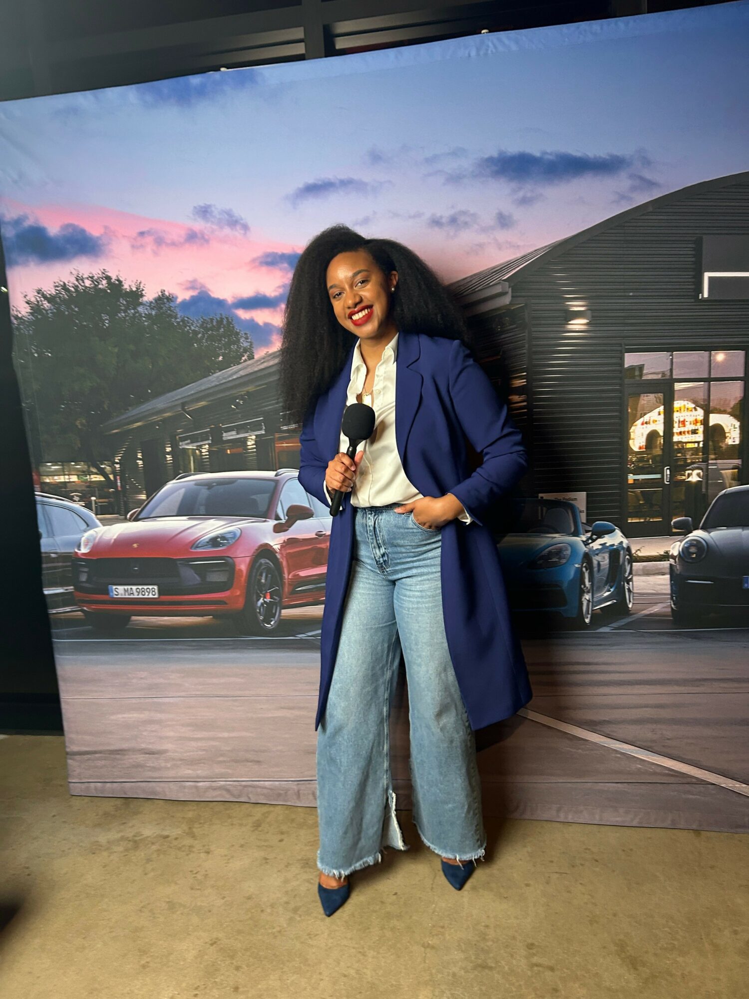 Woman standing in a parking lot at sunset, holding a phone, with cars and a building in the background.