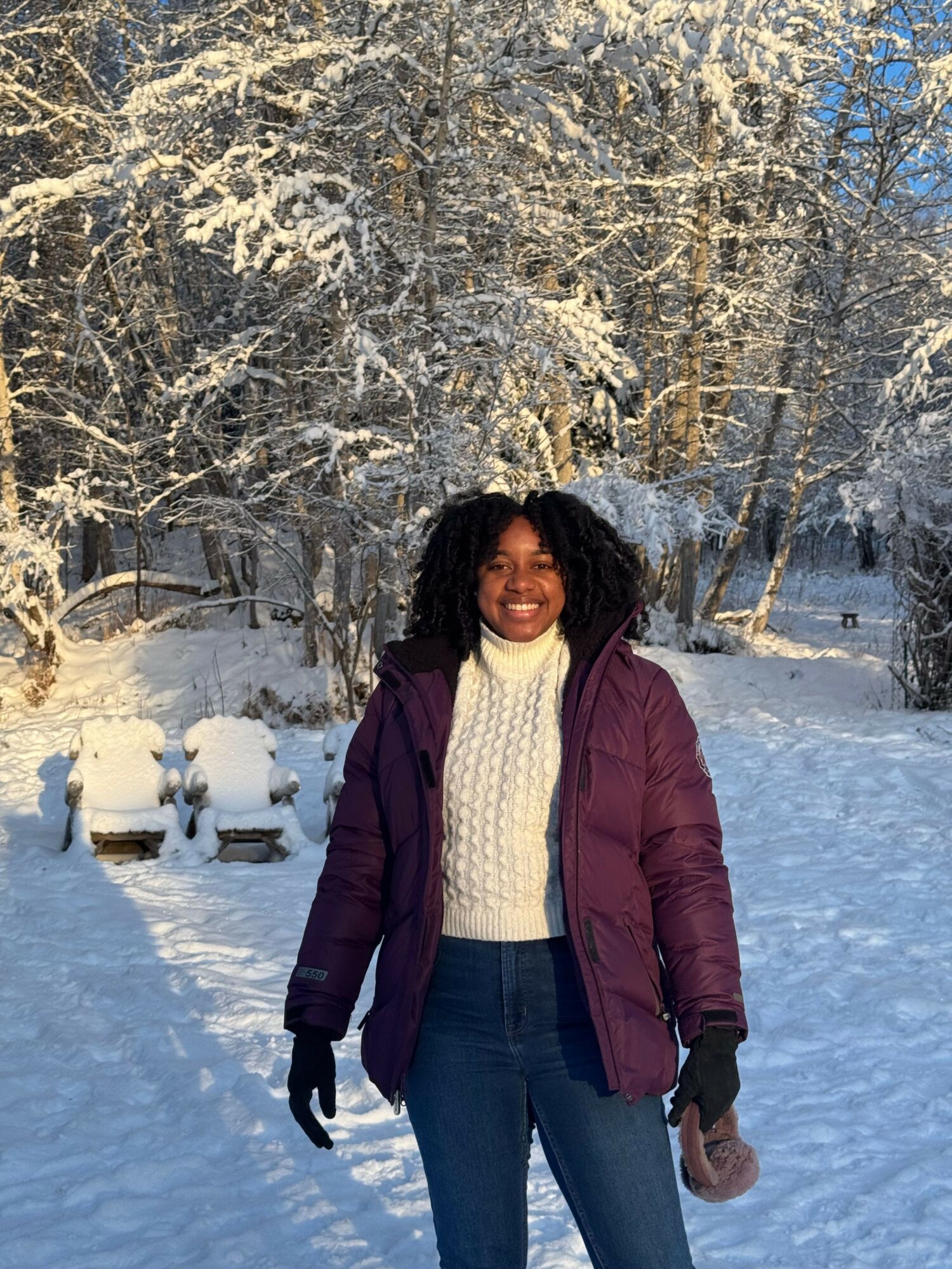 Woman standing outdoors in snow, wearing a maroon jacket, cream sweater, and black gloves, with snow-covered trees in background.
