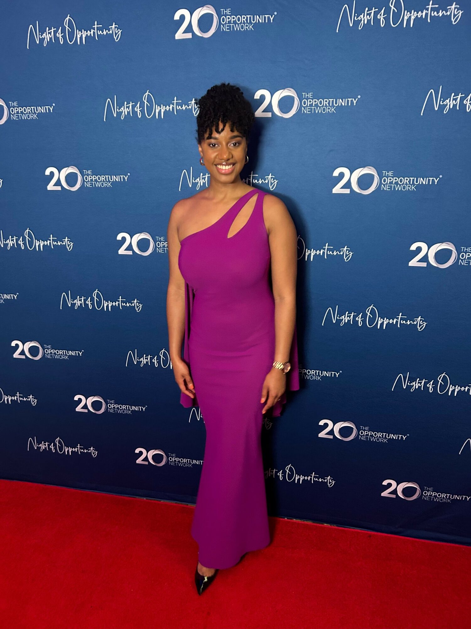 Woman in a purple dress standing on red carpet against blue backdrop with white text, posing for photo.