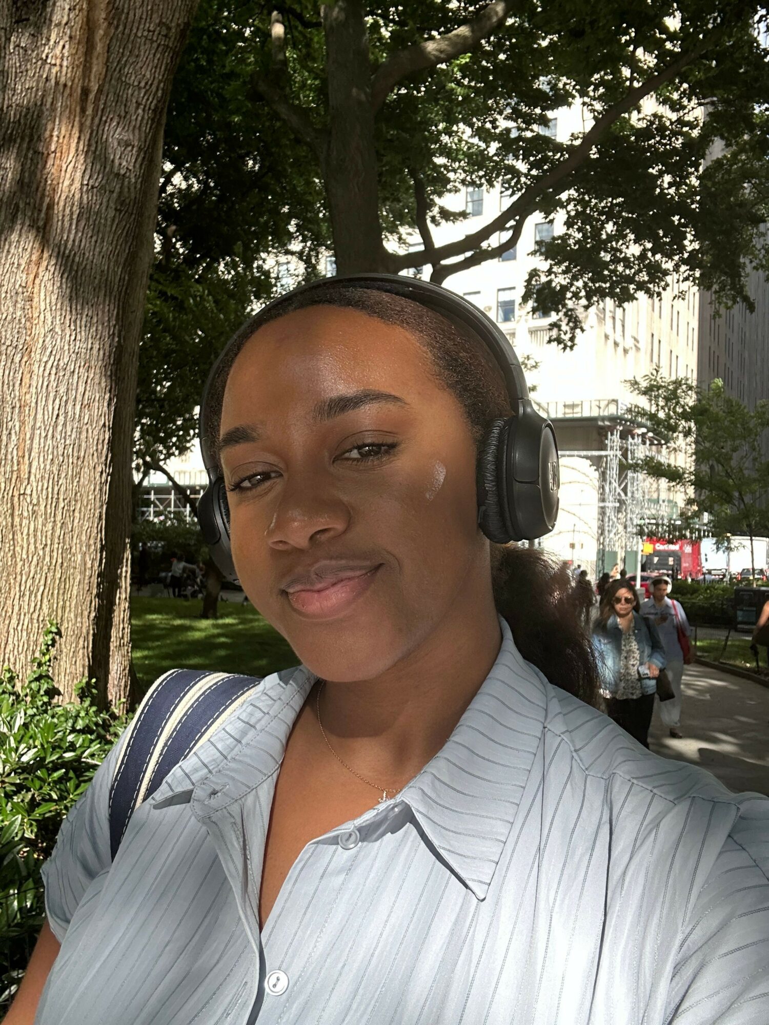 Woman with headphones outdoors, trees and park in background, smiling, wearing a light-colored shirt.