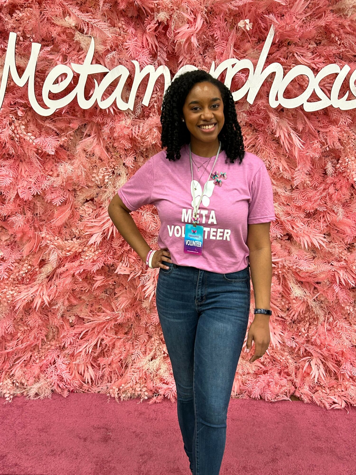 Woman standing in front of pink floral backdrop, wearing pink T-shirt and jeans, smiling, with text on backdrop and shirt.