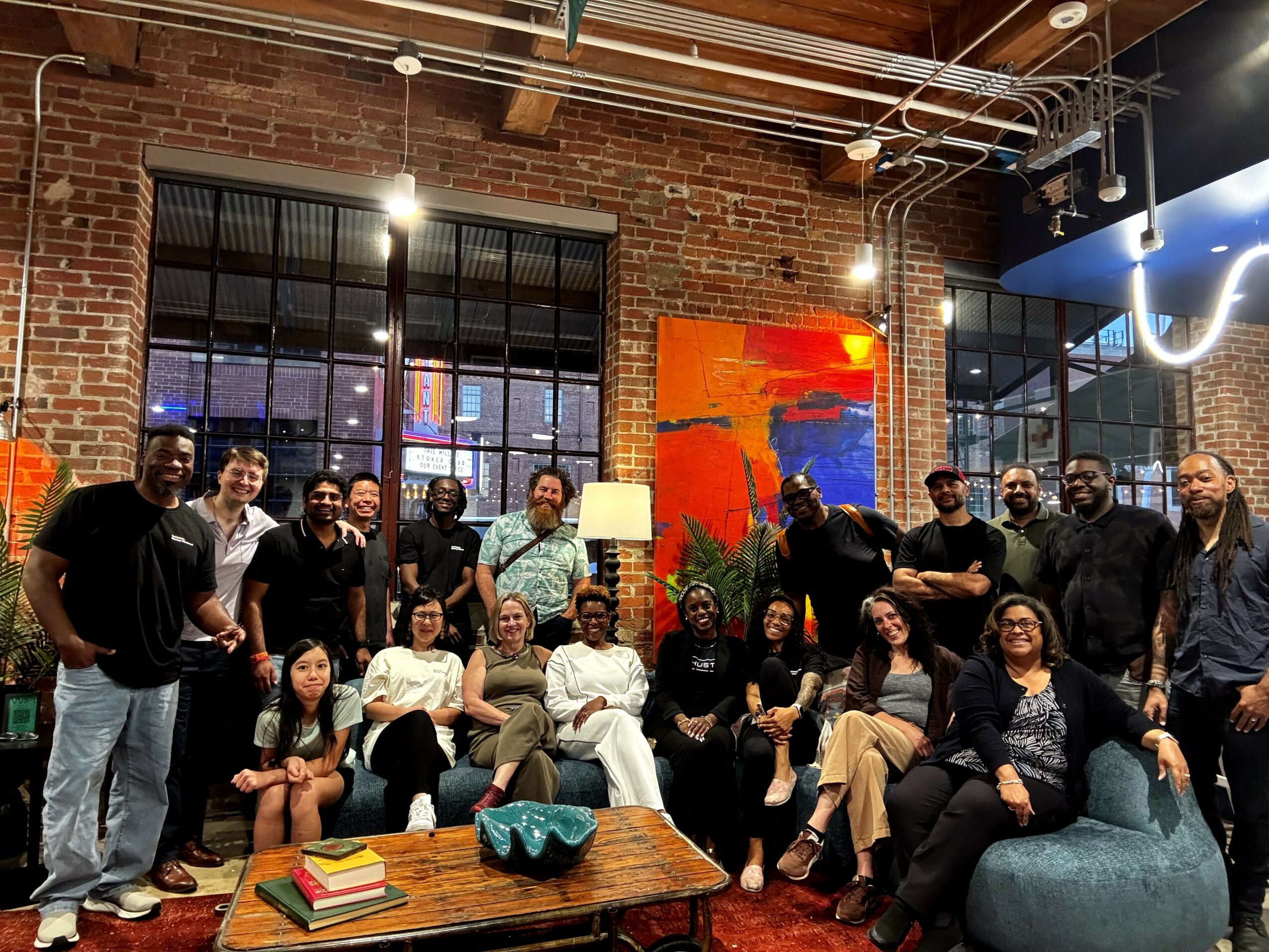 Group of people gathered in a brick-walled room with large windows, colorful artwork, and modern furniture, smiling and posing for a photo.