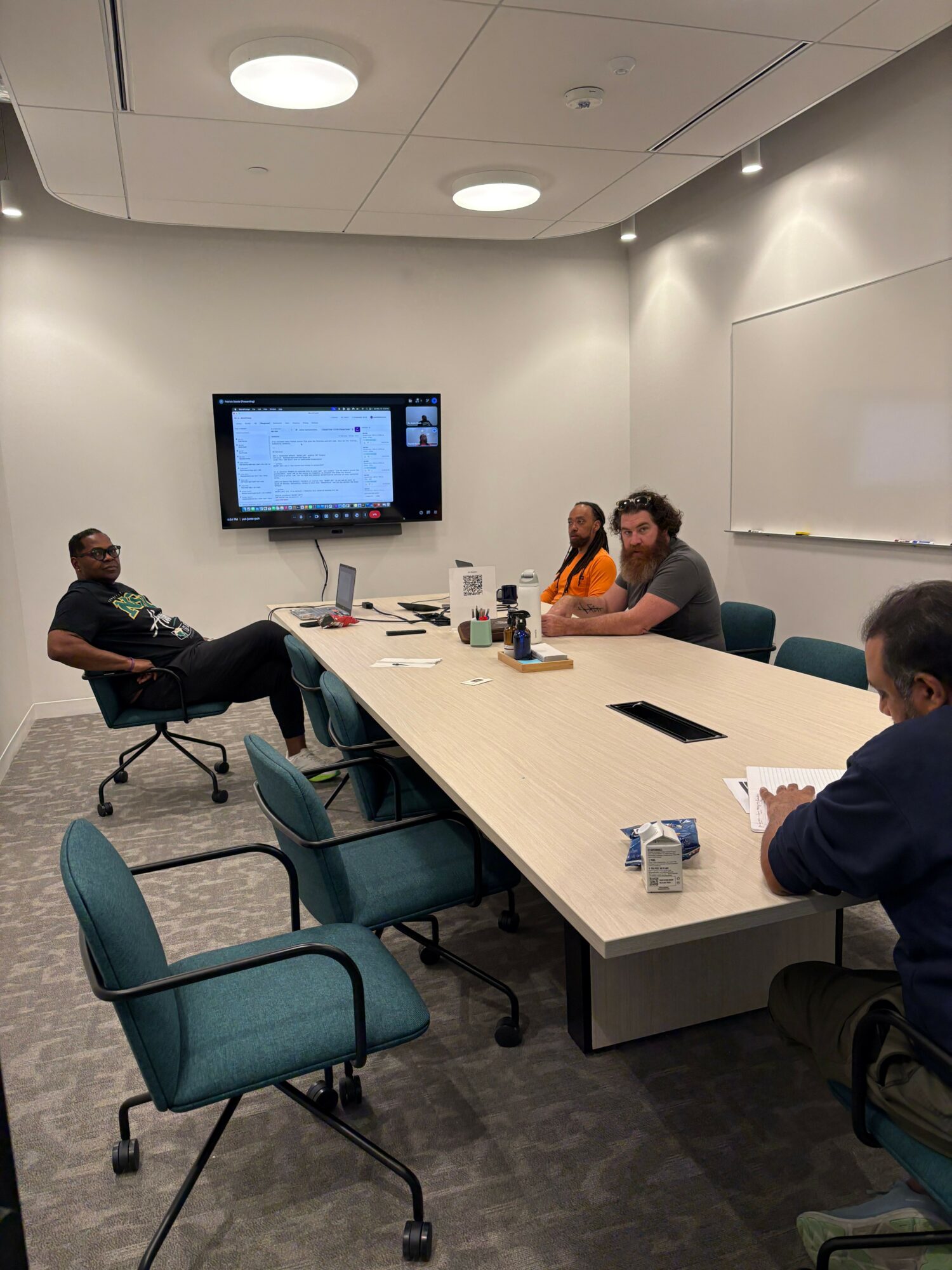 People sitting around a table with a monitor on the wall, papers, and drinks, in a conference room.