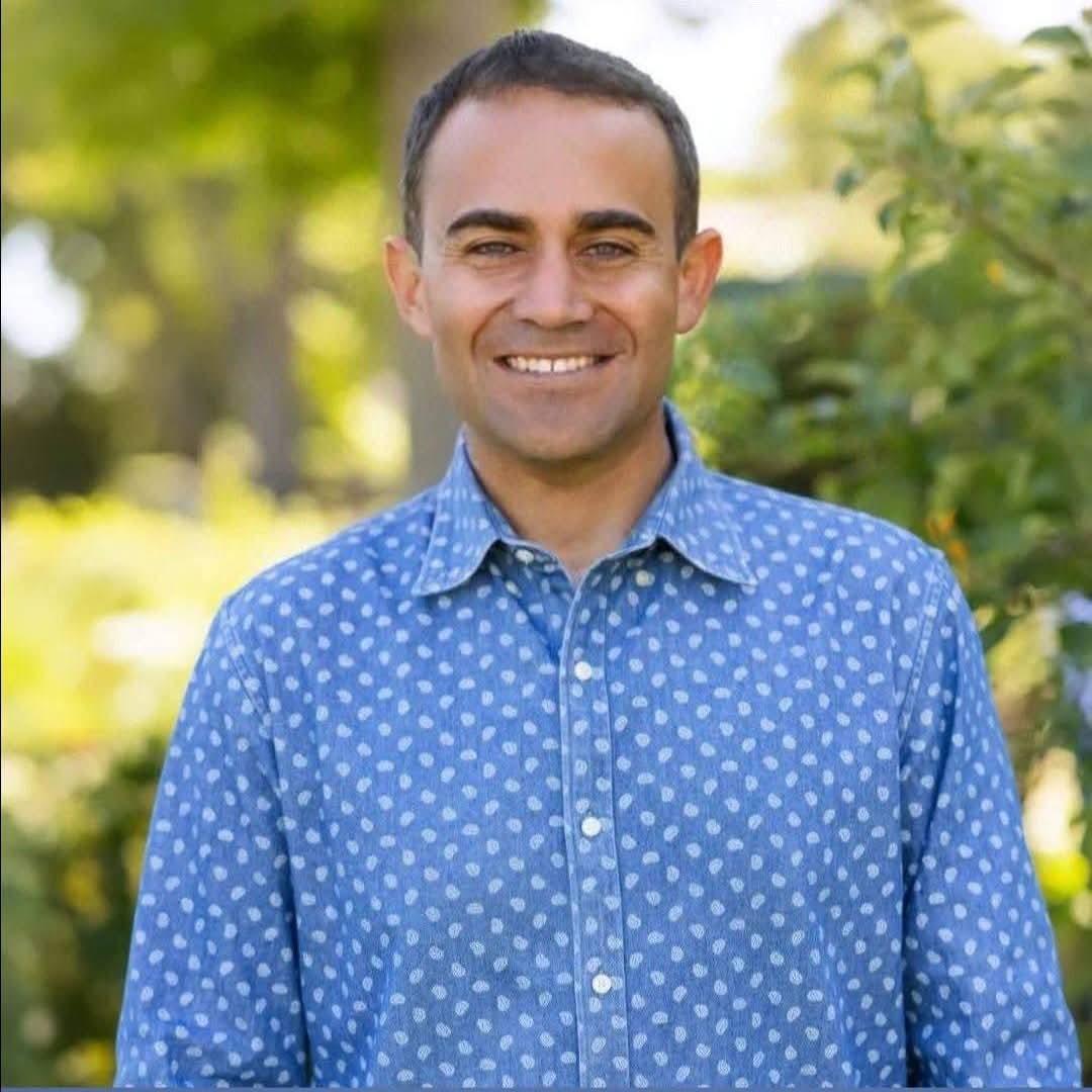 Man smiling outdoors with green foliage background, wearing a blue polka dot shirt.